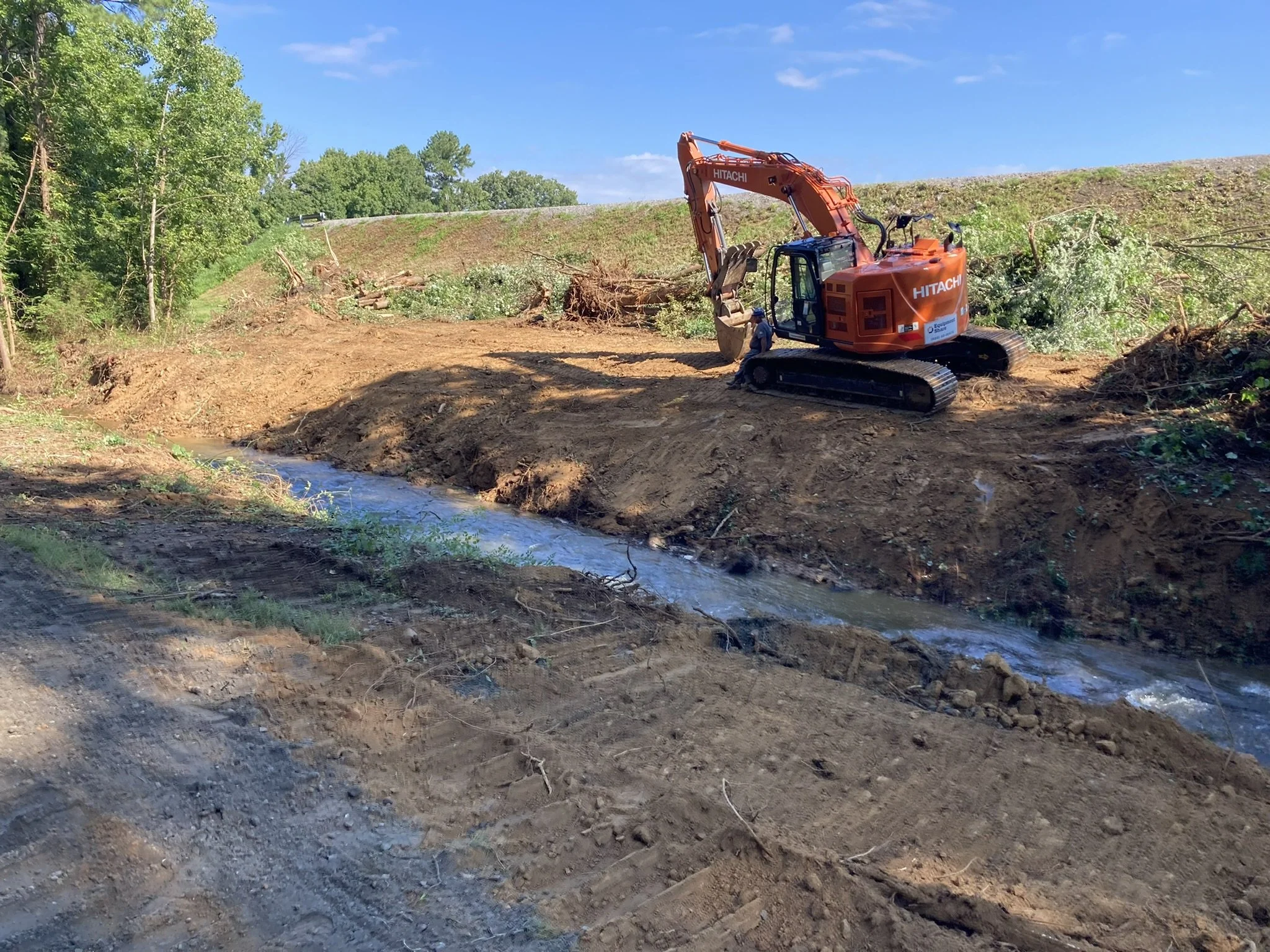 A construction site with an orange Hitachi excavator on a dirt embankment beside a small creek, surrounded by trees and a clear blue sky.