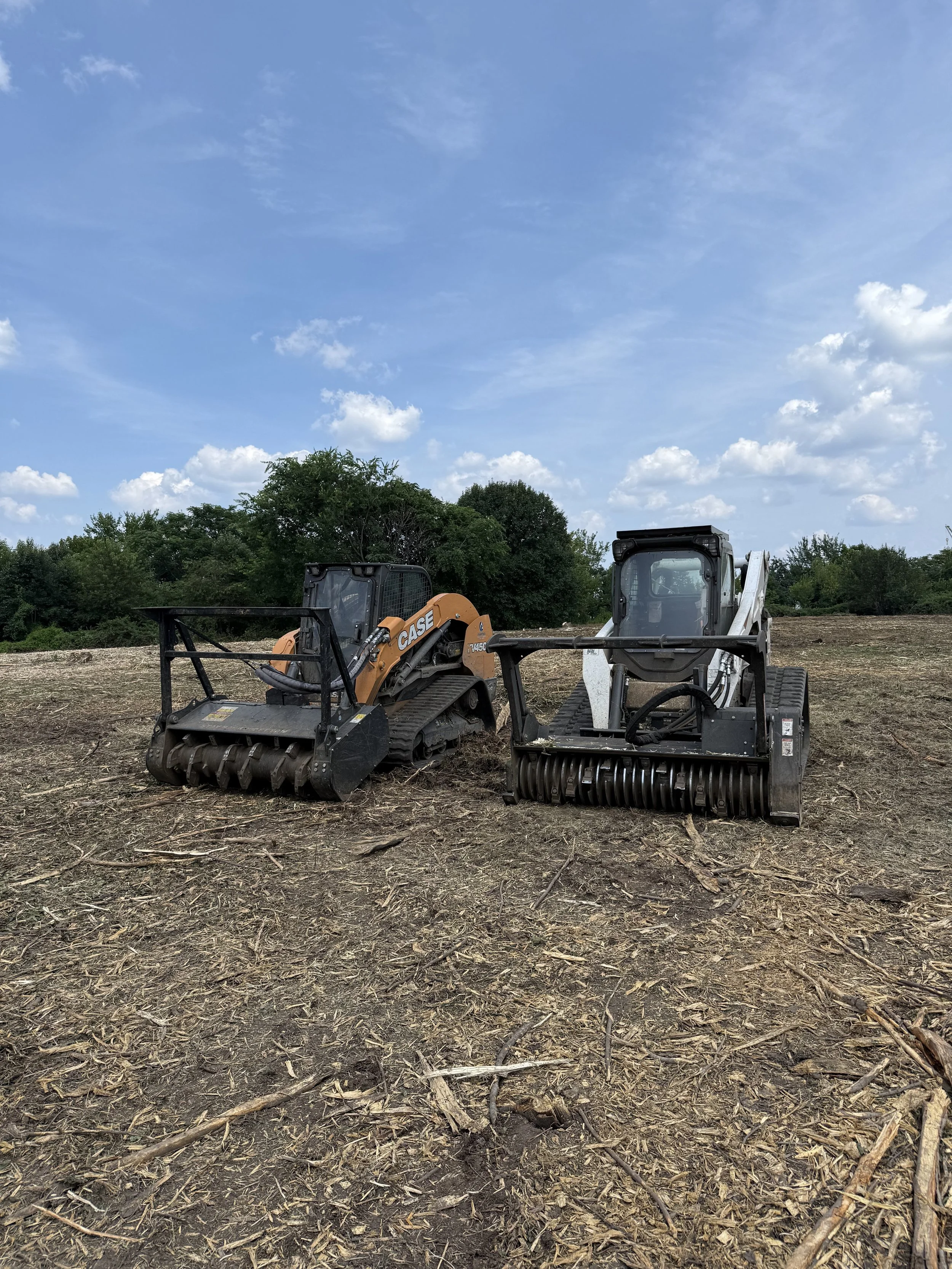 Two compact track loaders on a cleared dirt field under a partly cloudy sky with trees in the background.