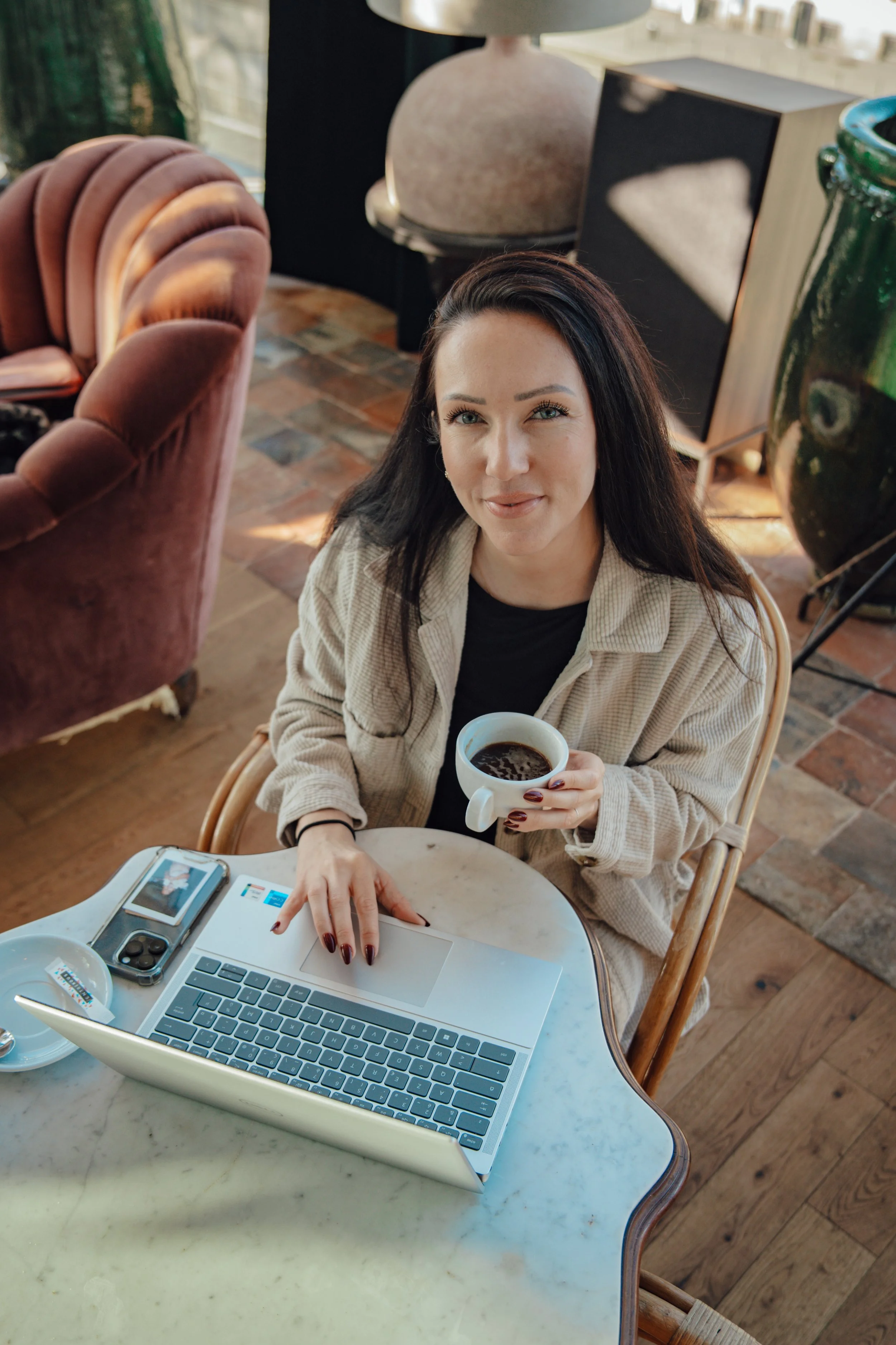 Une jeune femme avec les cheveux noirs, tenant une tasse de café, travaille sur un ordinateur portable blanc à une table en marbre dans un café.
