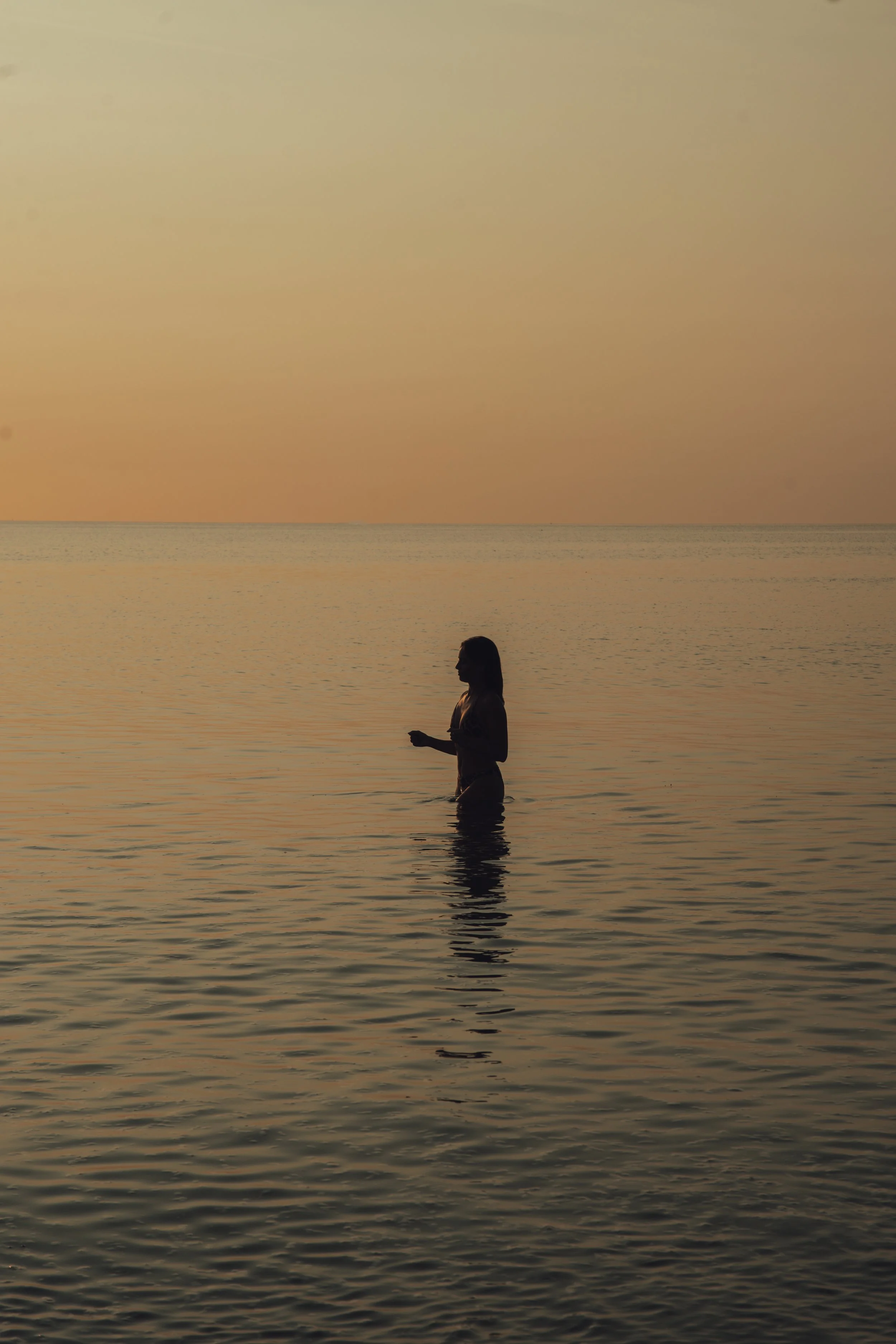Silhouette d'une femme dans l'eau au coucher du soleil, avec un ciel pastel et une mer calme.