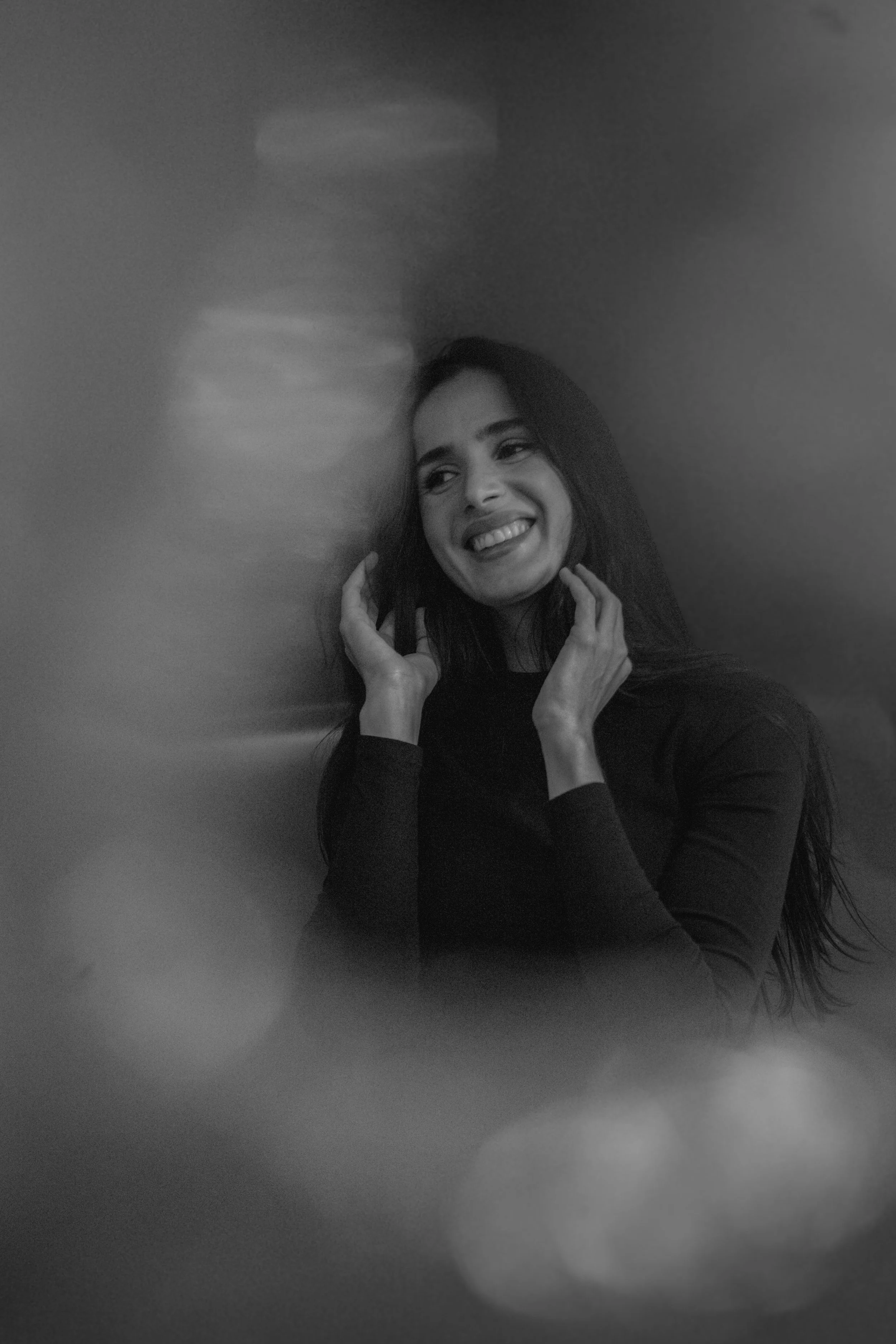Une femme souriante, en noir et blanc, posant contre un mur, entourée d'effets de flou lumineux.