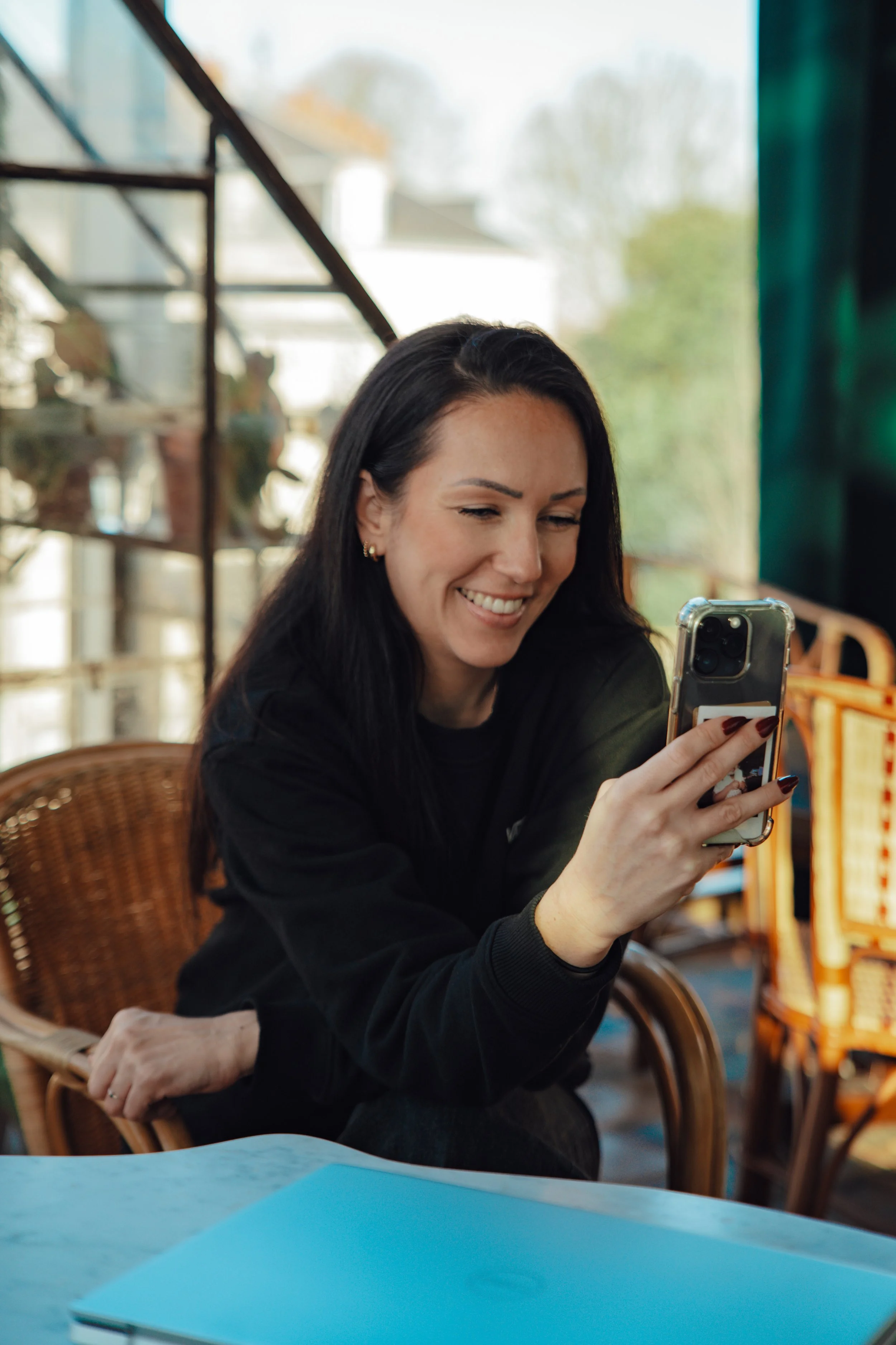 Une femme souriante regarde son téléphone, assise à une table dans un lieu lumineux avec des étagères en métal en arrière-plan.