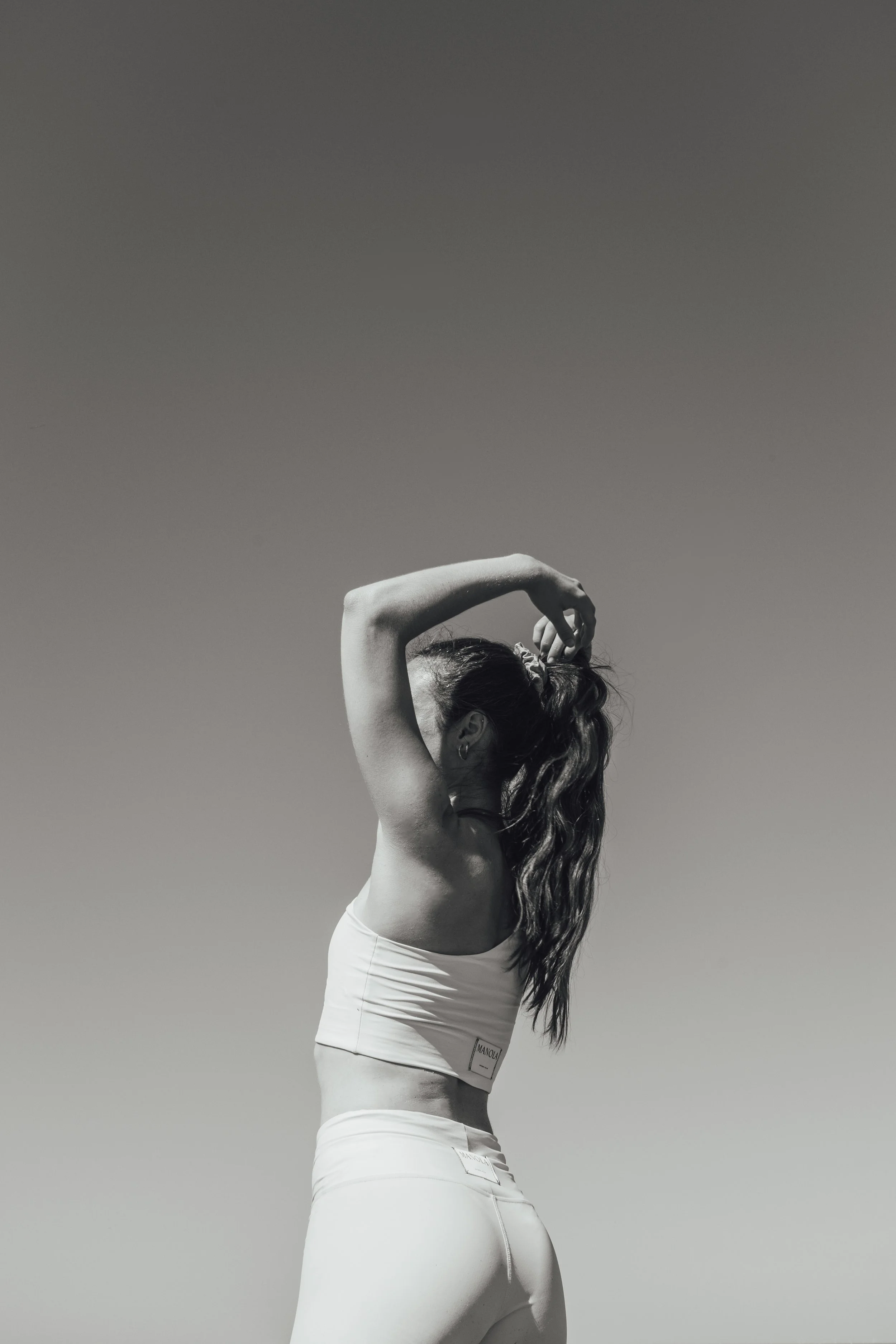 Une femme en vêtements de sport, vue de profil, qui ajuste ses cheveux avec un bras levé, en plein air contre un ciel clair.