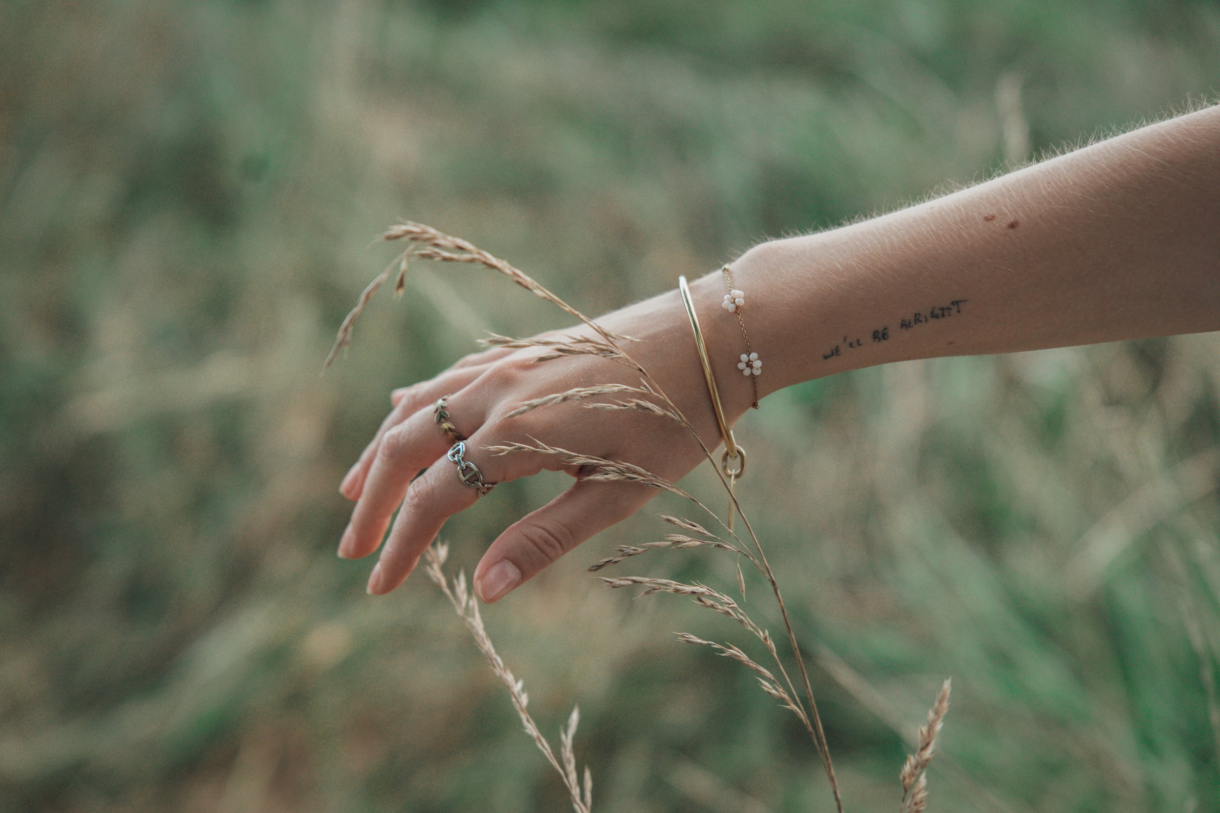 Une main féminine portant plusieurs bijoux,main tachetée avec un tatouage en français, dans un champ d'herbes grasses