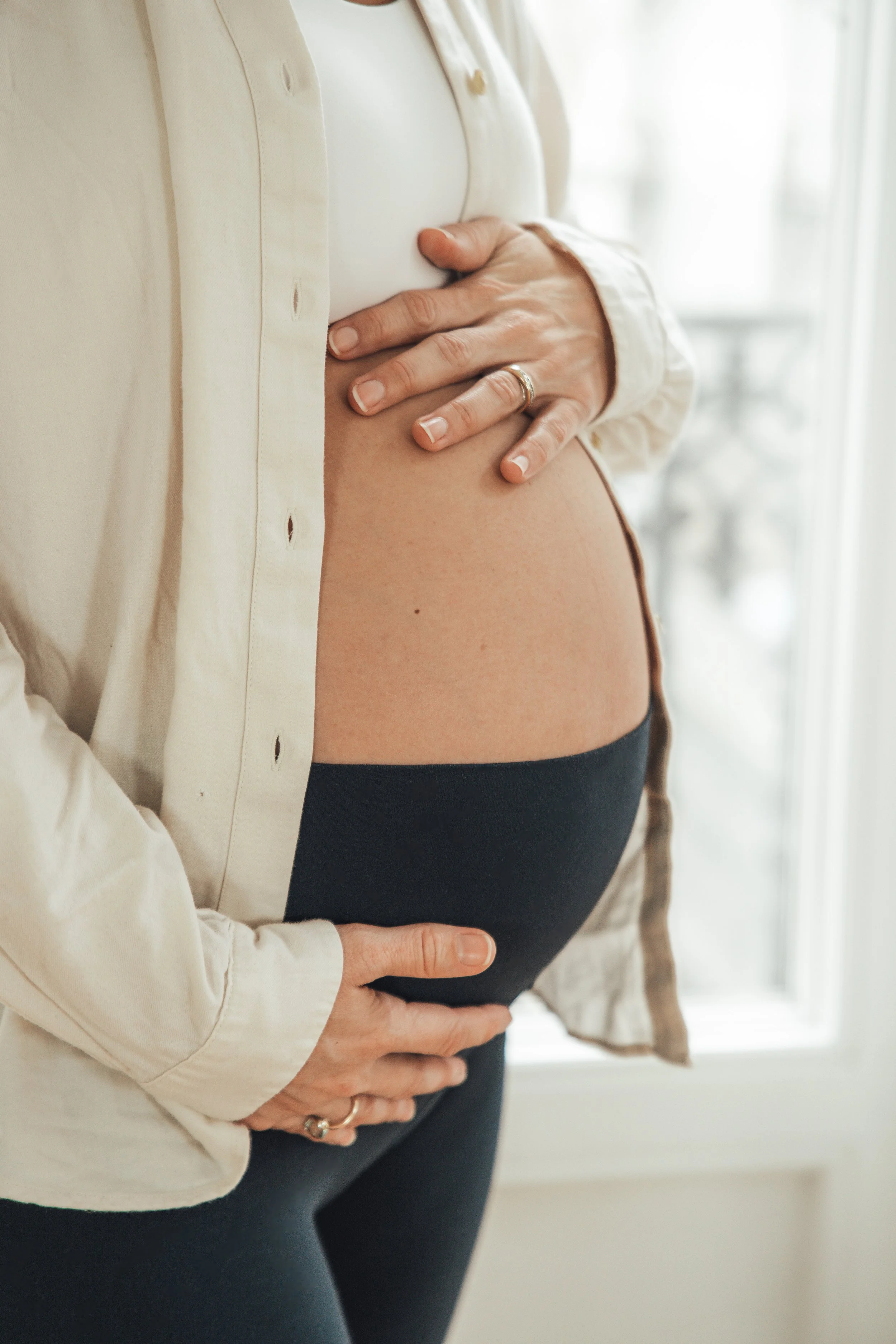 Une femme enceinte tient son ventre rond avec ses mains, près d'une fenêtre, dans un intérieur lumineux.