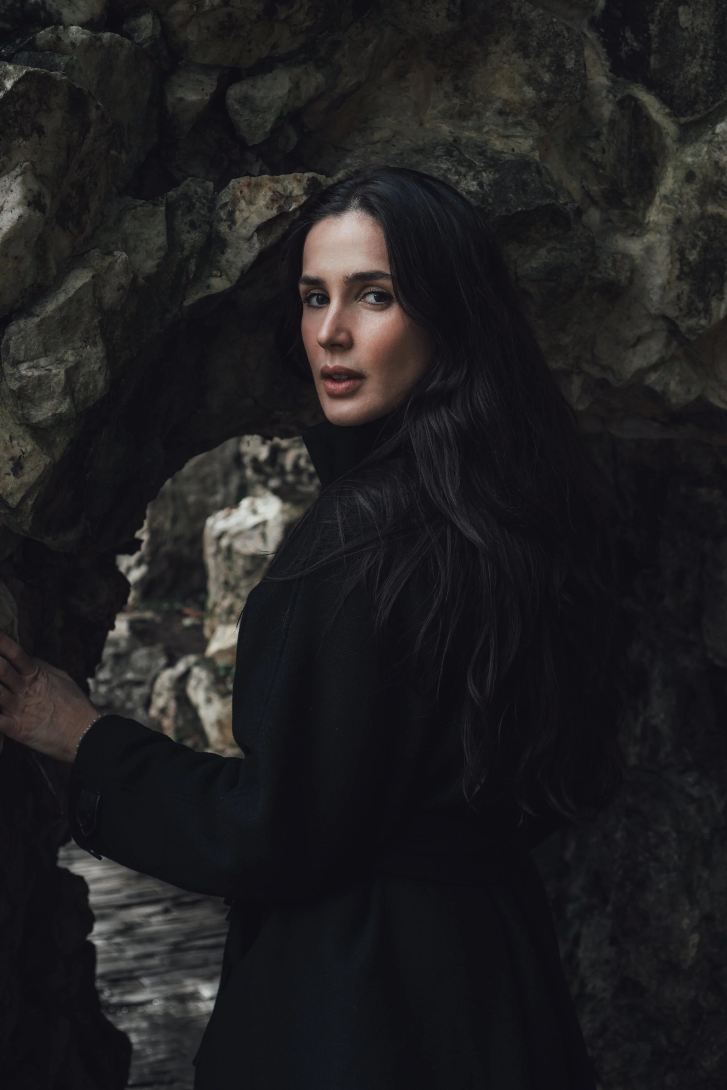 Femme aux cheveux longs et noirs, portant un manteau noir, se tient près d'une structure rocheuse, dans un environnement naturel.