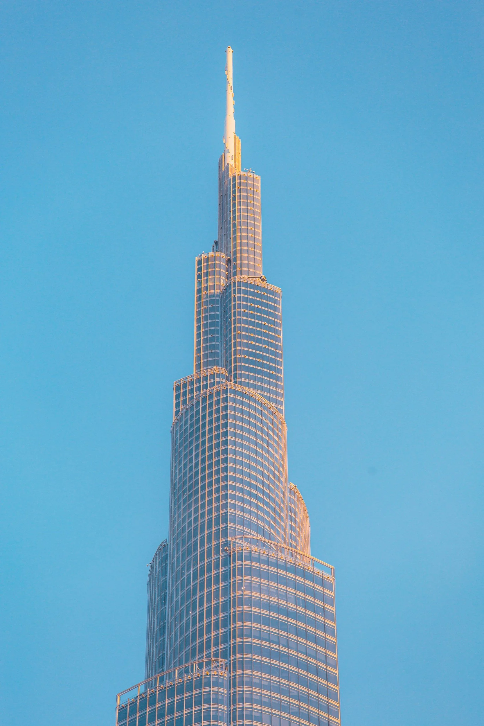 The tall, glass skyscraper with tiered sections rises into a clear blue sky, bathed in warm sunlight.