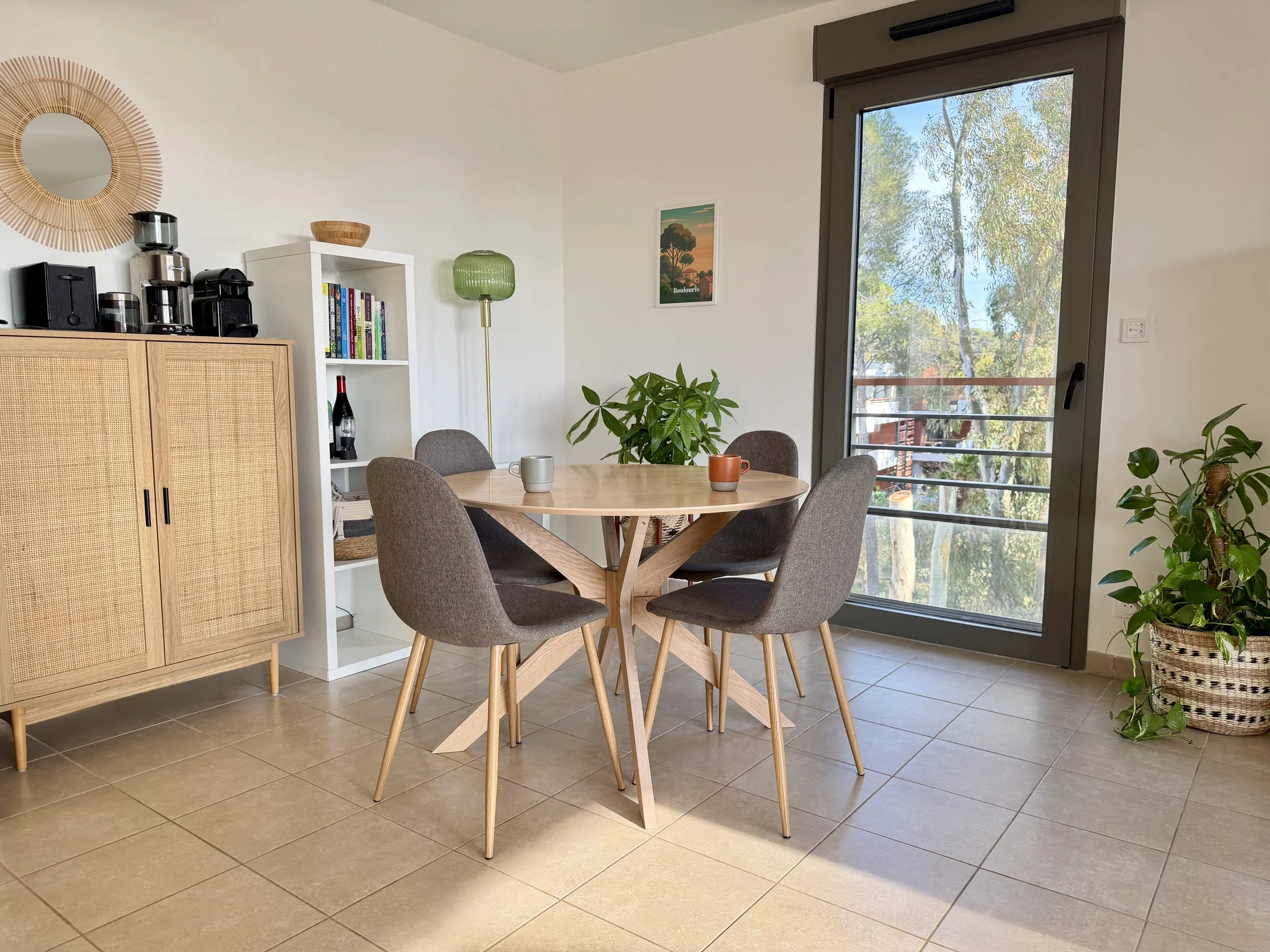 Salle à manger avec une table en bois, quatre chaises en tissu gris, une grande fenêtre avec un balcon, une plante verte en pot, une étagère blanche avec des livres, un lampadaire vert, un meuble en bois avec appareils électroménagers et une décoration en miroir en rotin