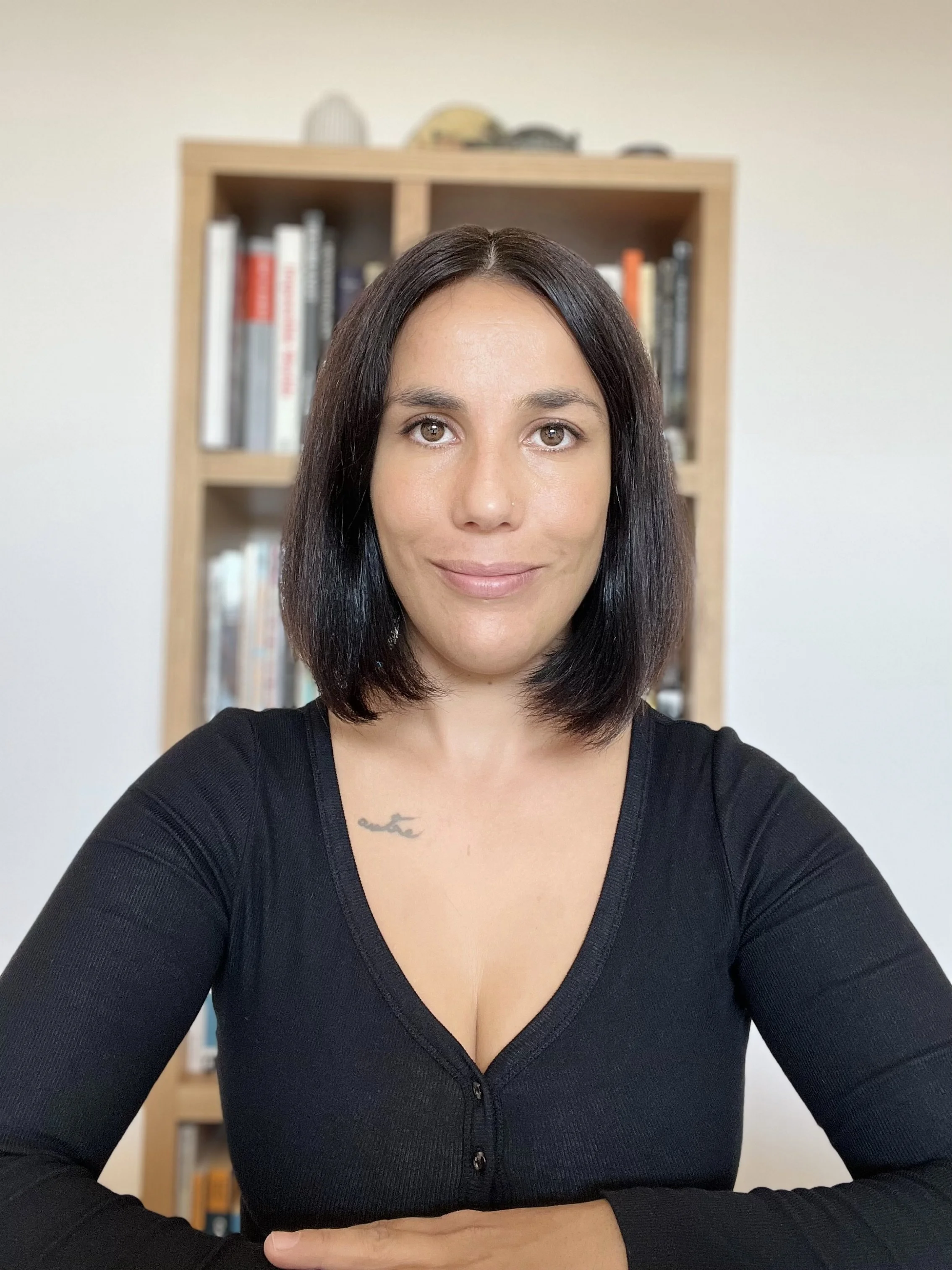 Une femme souriante avec les cheveux courts et noirs. Elle porte un haut noir à boutons. Elle est assise devant une étagère en bois contenant des livres et des décorations.