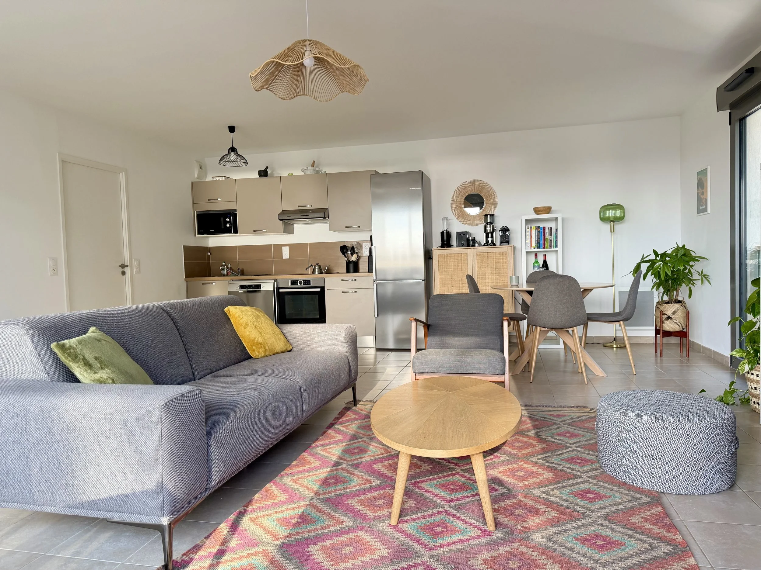 Salon et cuisine moderne avec sofa gris, table basse ronde en bois, pouf gris à motifs, table à manger avec 4 chaises, plantes vertes, luminaires suspendus et décorations