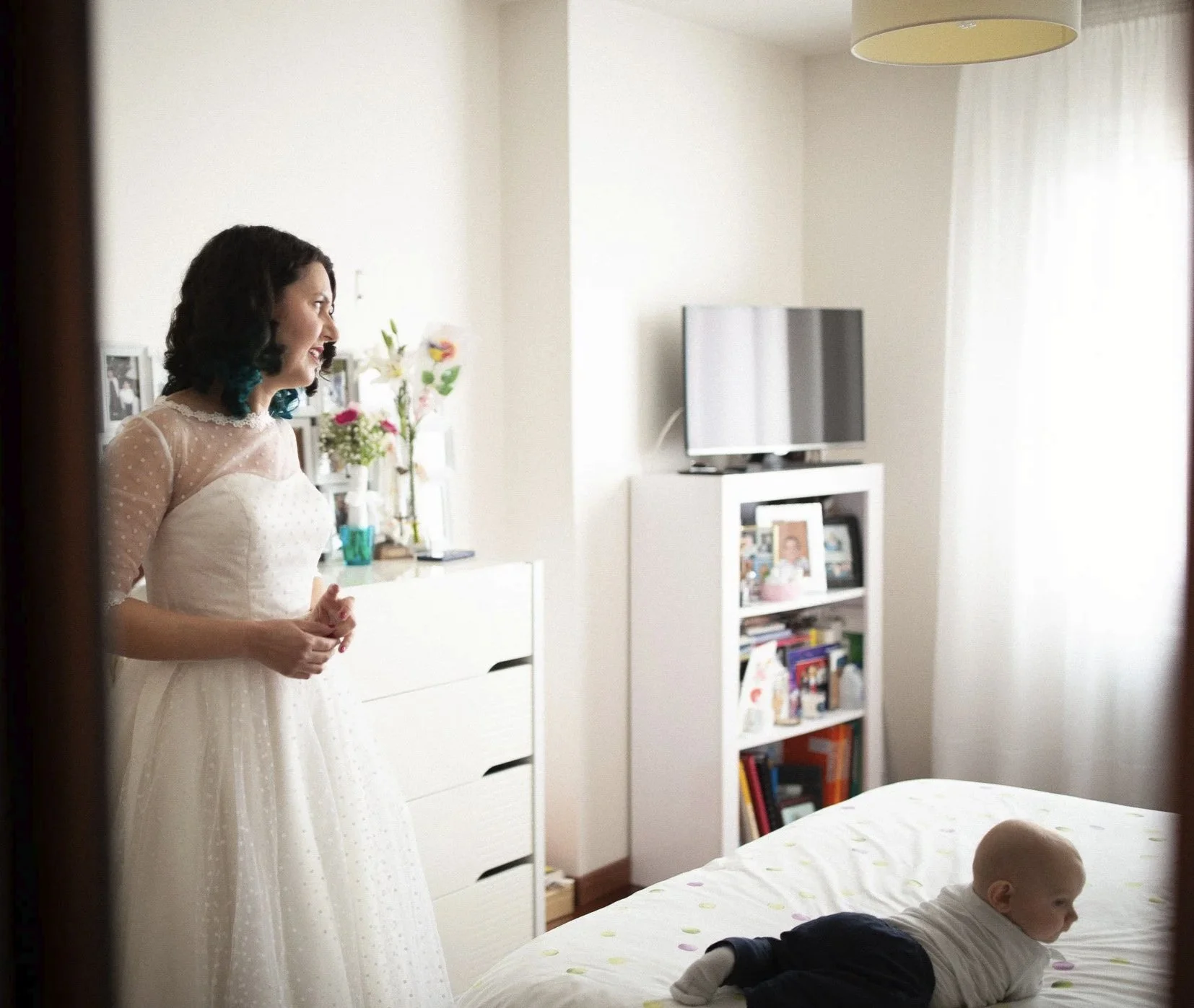 Une femme en robe blanche souriante observant un bébé allongé sur un lit dans une chambre lumineuse avec des meubles et décorations pendant le reportage photo d'un mariage.