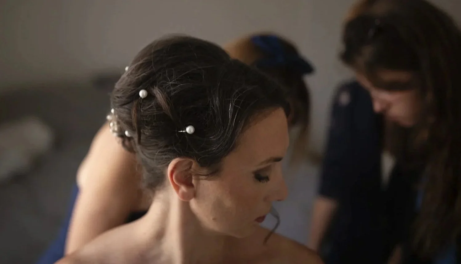 Une femme avec des cheveux bruns ornés de perles, probablement une mariée, reçoit de l'aide pour sa coiffure par une autre femme lors de préparatifs de son mariage.