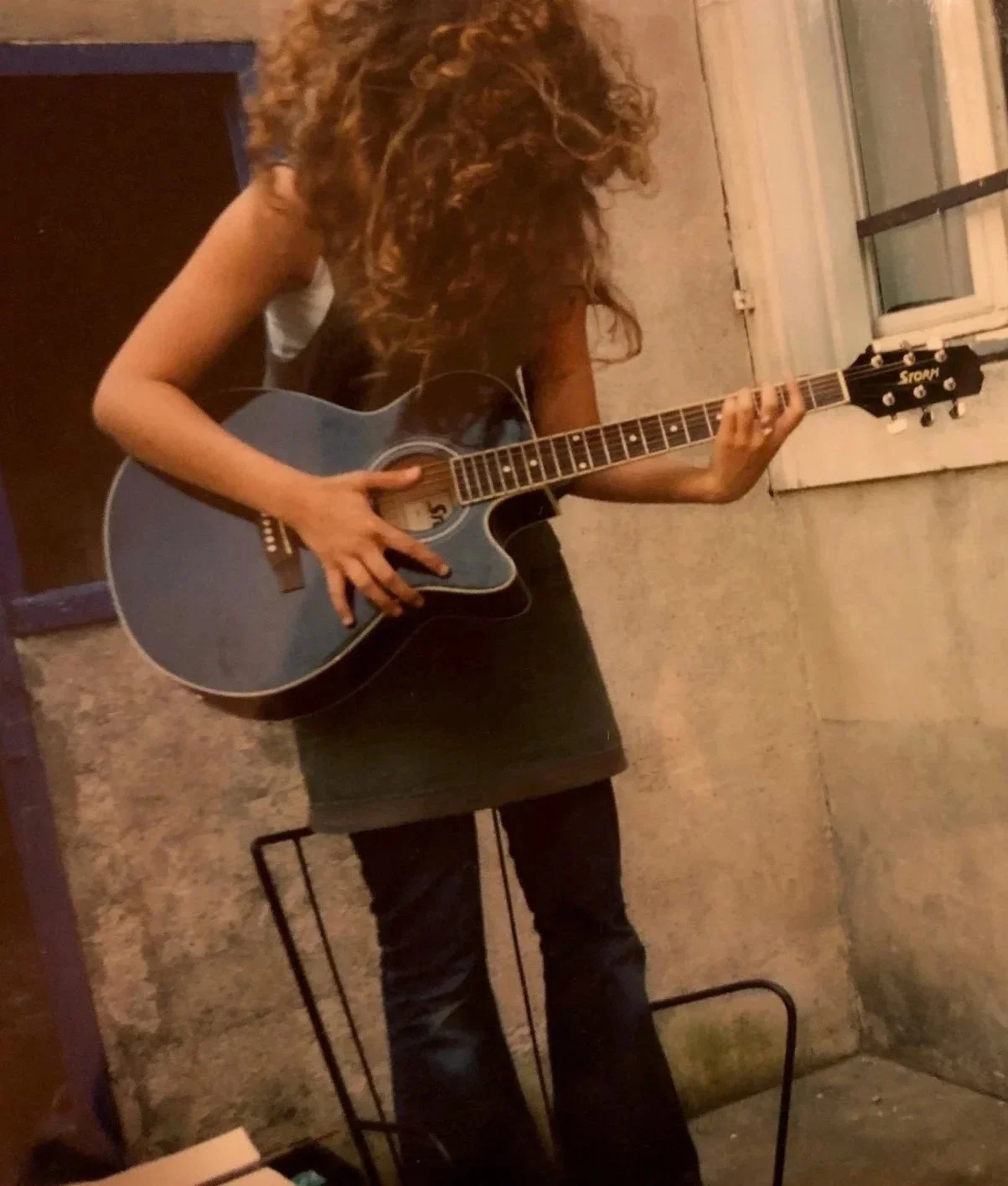 Une femme joue de la guitare acoustique dans une pièce avec des murs en béton et une fenêtre