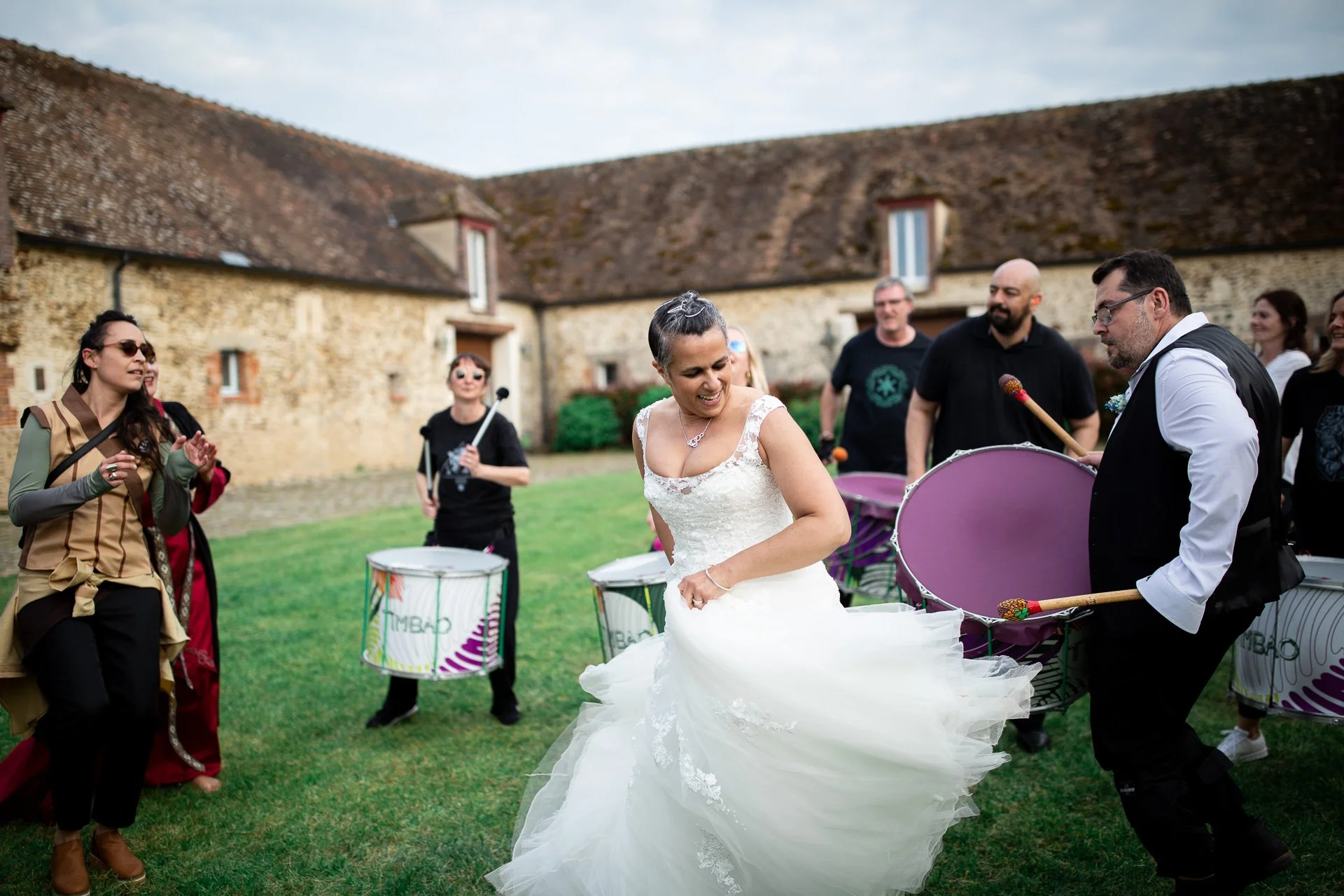 Un groupe de personnes joue des tambours lors d'un mariage en plein air, une femme en robe de mariage danse au centre, pendant un reportage photo de mariage en ile de France.