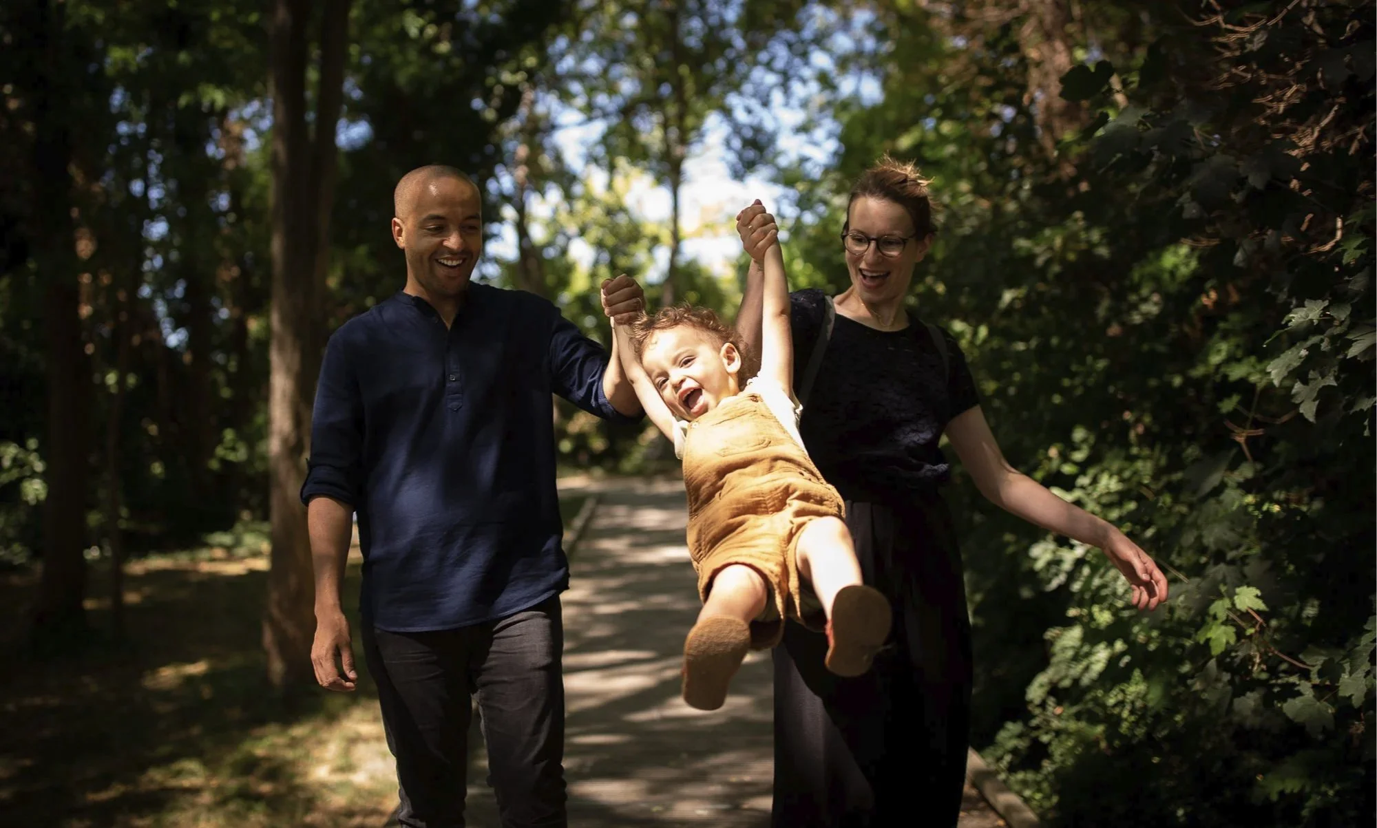 Une famille marche dans un parc à Montreuil en profitant d'une journée ensoleillée, avec un enfant joyeux suspendu lors d'une séance photo de famille.