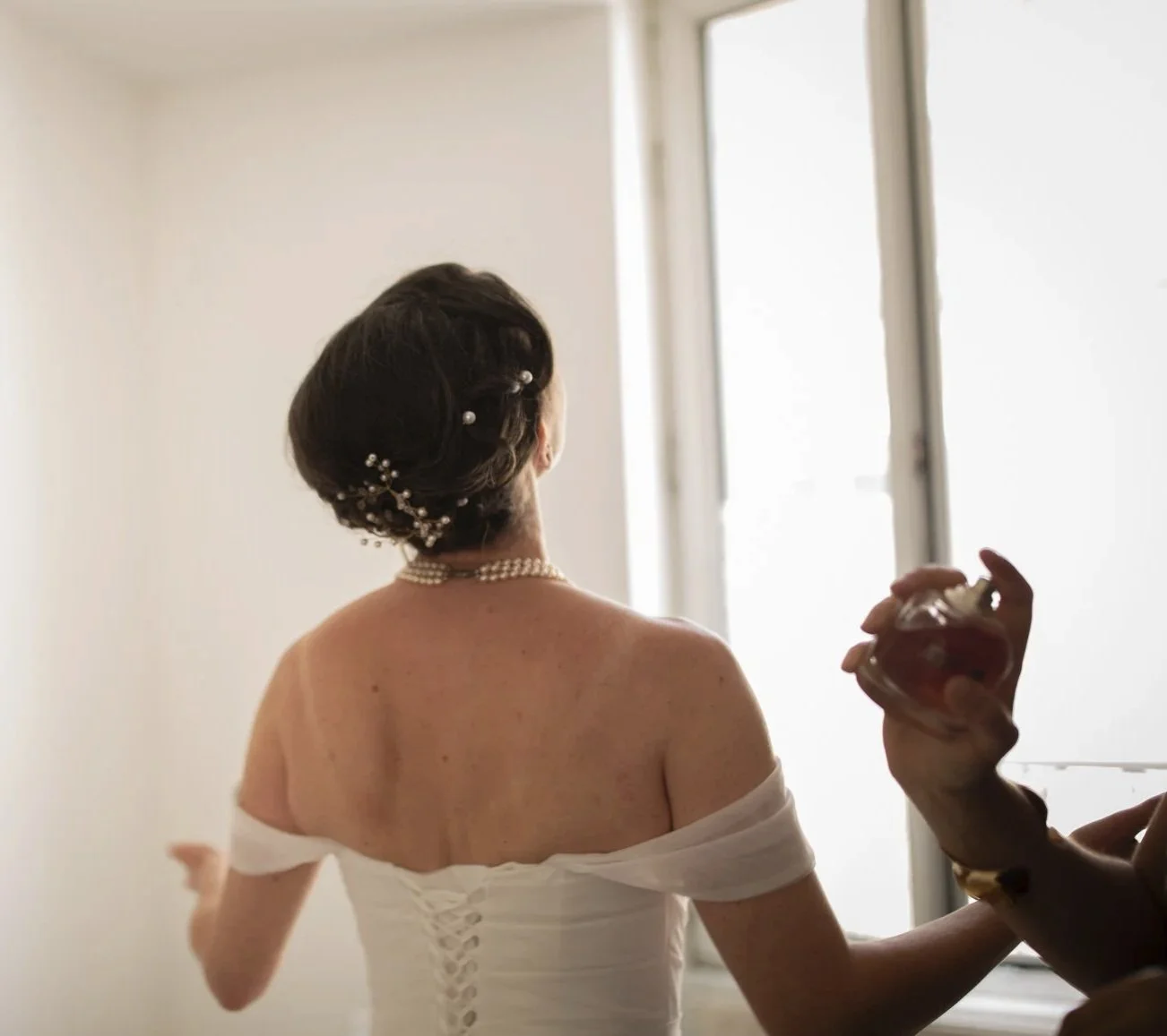 Une femme en robe blanche se prépare, probablement pour un mariage, avec une coiffure sophistiquée décorée de perles, portant un collier de perles, devant une fenêtre lumineuse pendant un reportage photo de mariage à Paris.