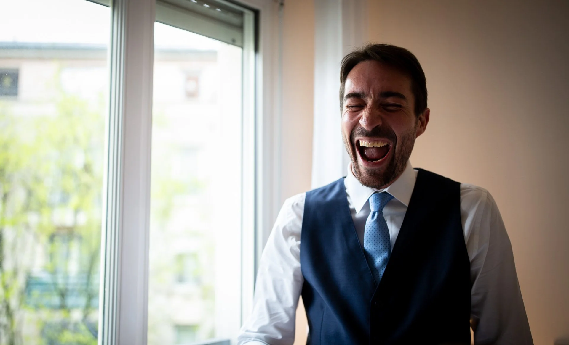 Un homme souriant et riant, portant une chemise blanche, un nœud papillon bleu, et un veston noir, debout près d'une fenêtre pris en photo lors des préparatifs de son mariage.