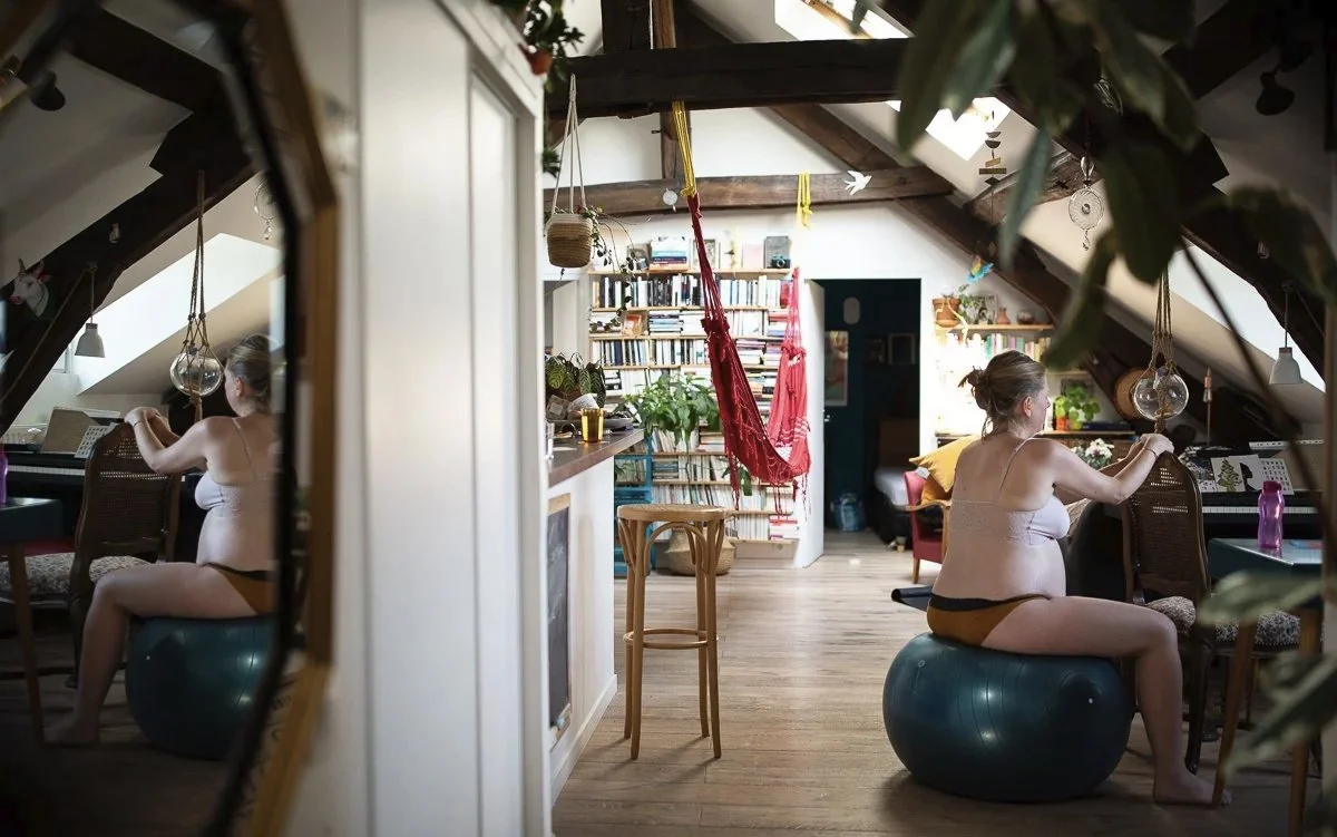 Une femme nue assise sur un ballon de grossesse à son domicile en travail avant son accouchement, pendant un reportage photo d'accouchement à Paris.
