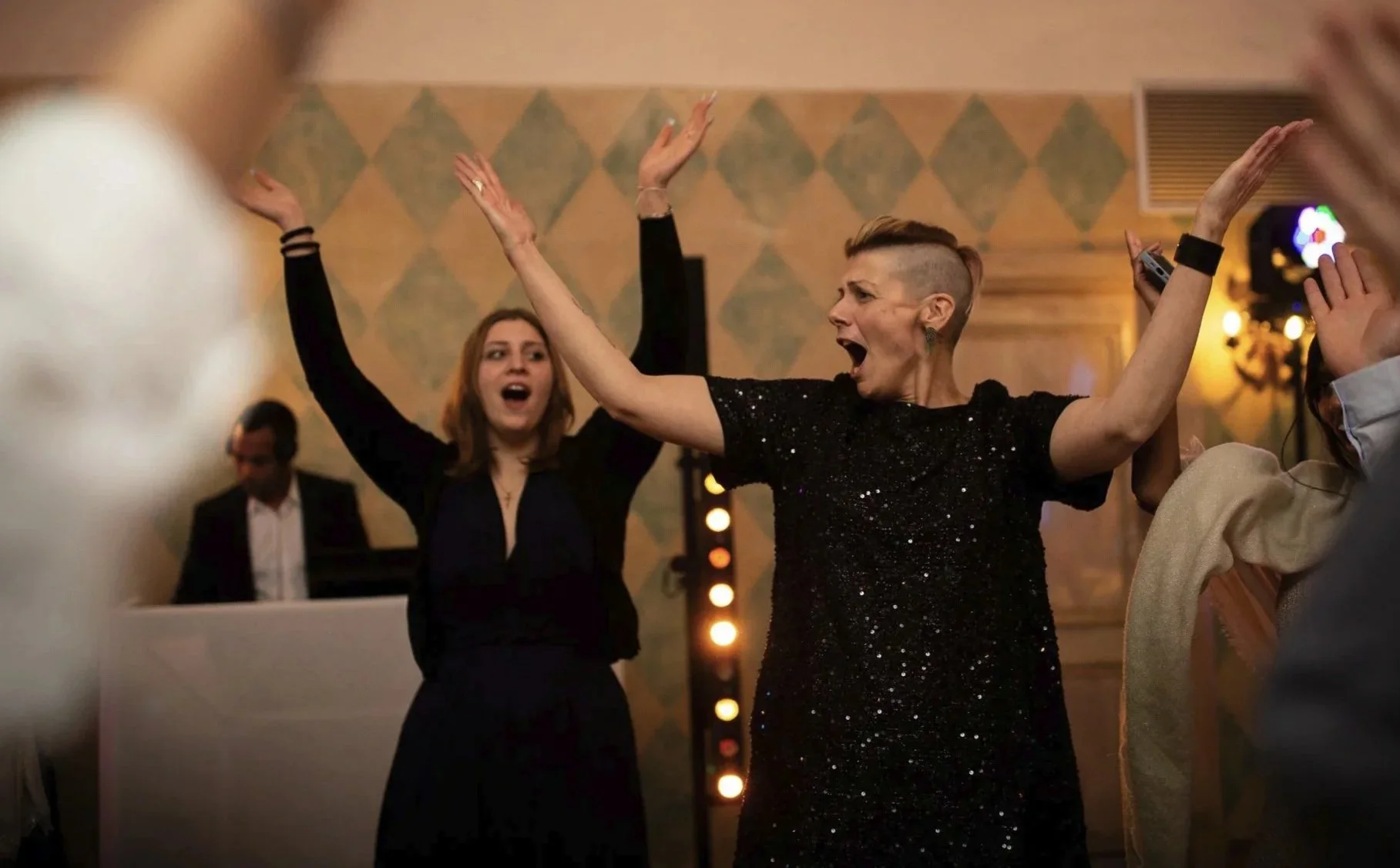Deux femmes en train de danser et de chanter lors d'une fête de mariage dans une salle élégante, avec un fond de mur décoré et un DJ à gauche pendant un reportage de mariage en Ile de France.