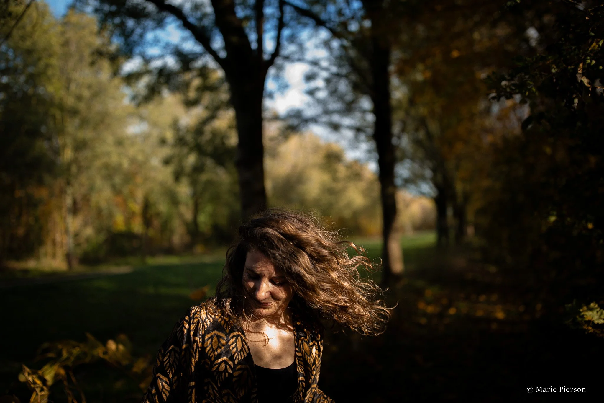 Une femme avec de longs cheveux bouclés et vêtue d'une robe à motifs dorés, dans un parc avec des arbres aux feuilles d'automne, sous une lumière douce lors d'une séance photo documentaire.