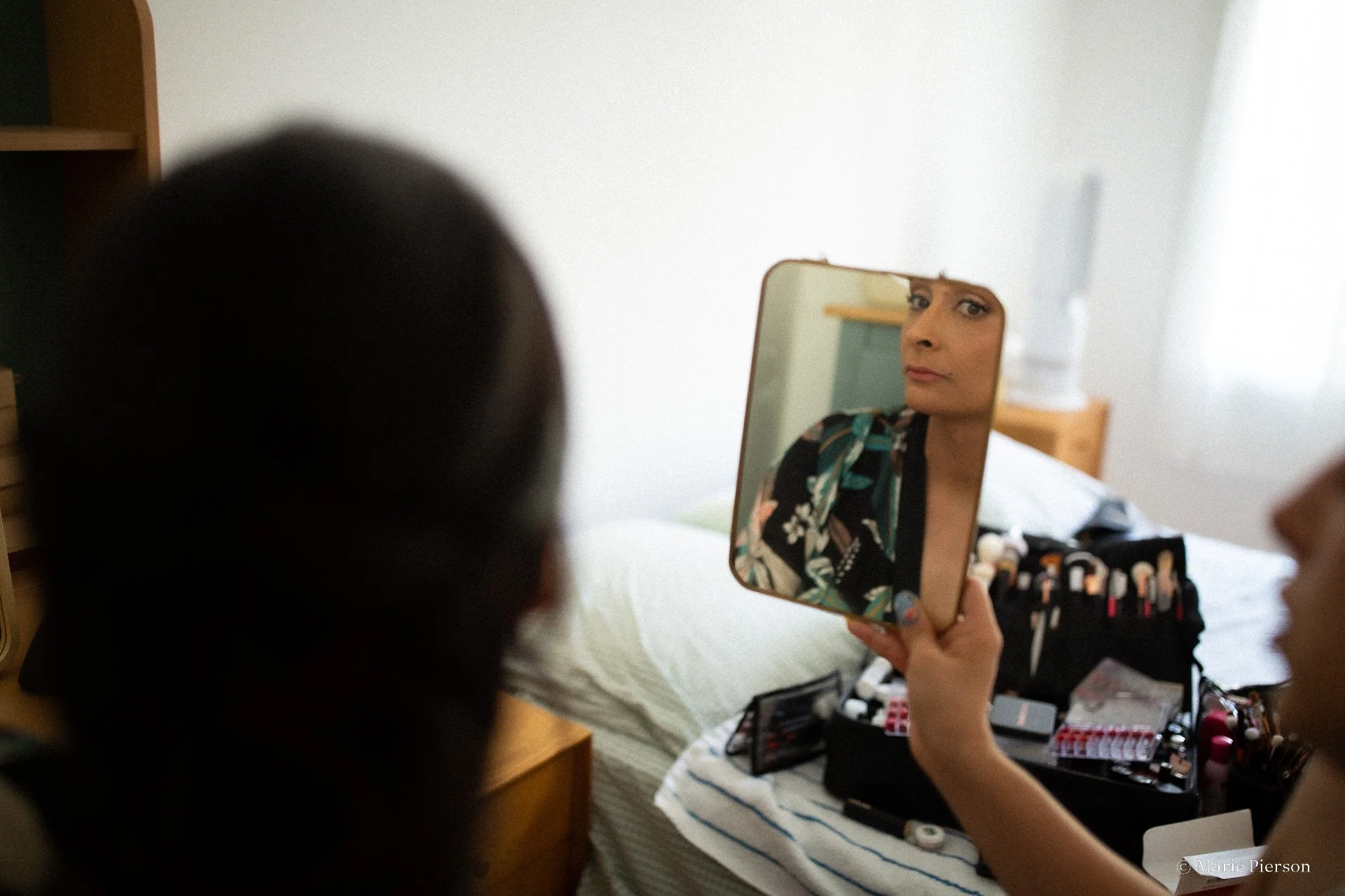 Une femme se prépare pour son mariage et regarde son reflet dans un miroir portable tout en appliquant du maquillage ou un gloss sur ses lèvres, dans une chambre.