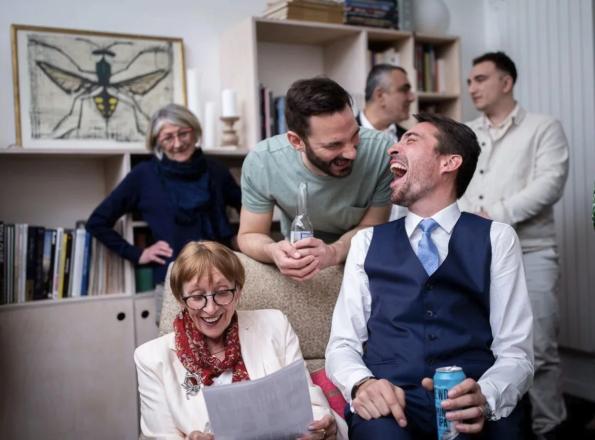 Groupe de six personnes souriantes et riant lors d'un cocktail de mariage pris en photo dans un salon, avec des livres et une peinture d'insecte en arrière-plan.