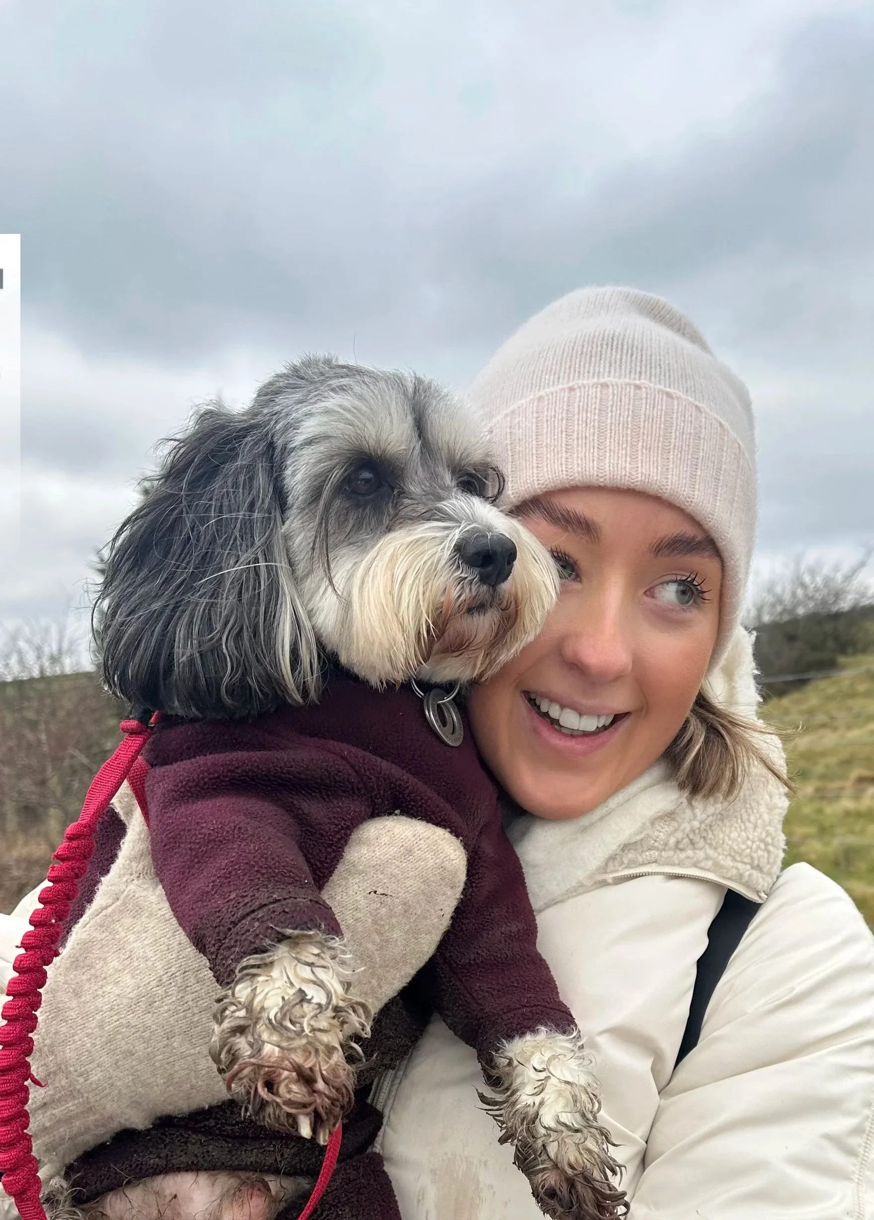A woman wearing a beige beanie and white jacket holding a small, fluffy dog with black, white, and gray fur, dressed in a maroon coat outdoors on a cloudy day.