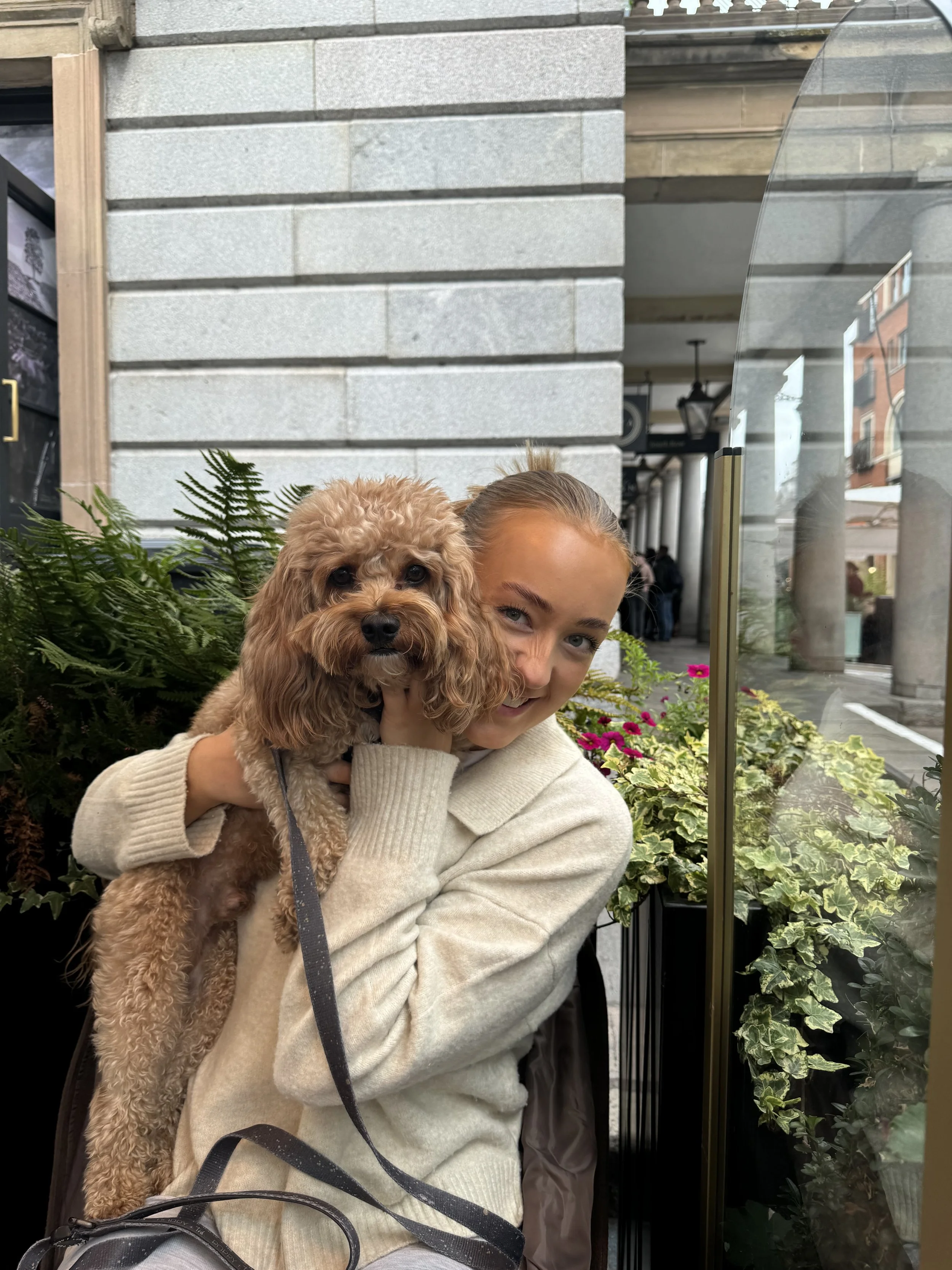 A young woman holding a small, curly-haired brown dog outside a building with plants and flowers nearby.