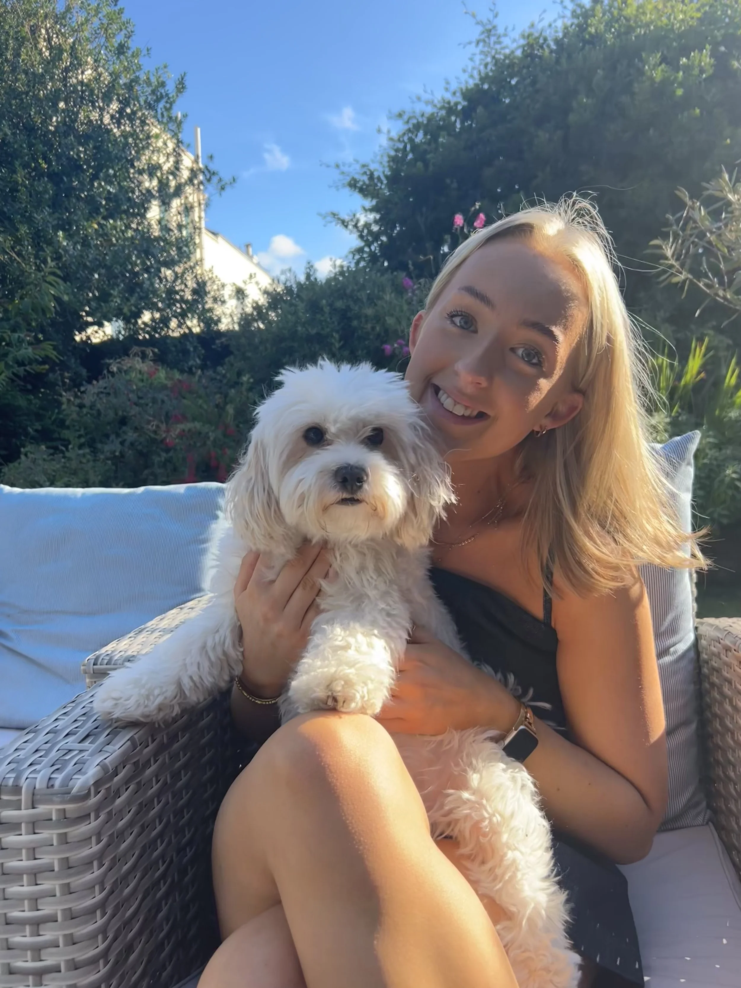 A woman sitting outdoors on a wicker couch, smiling and holding a small white fluffy dog, with green bushes and trees in the background on a sunny day.