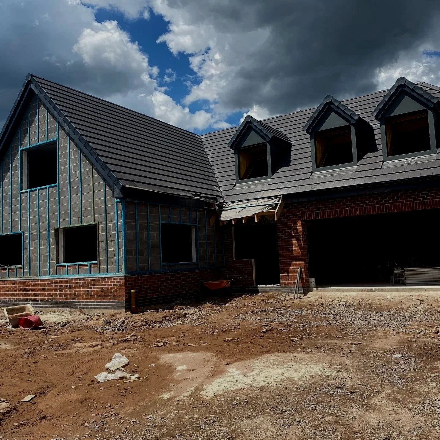 Construction of a house with brick walls and a gray shingle roof under a partly cloudy sky.