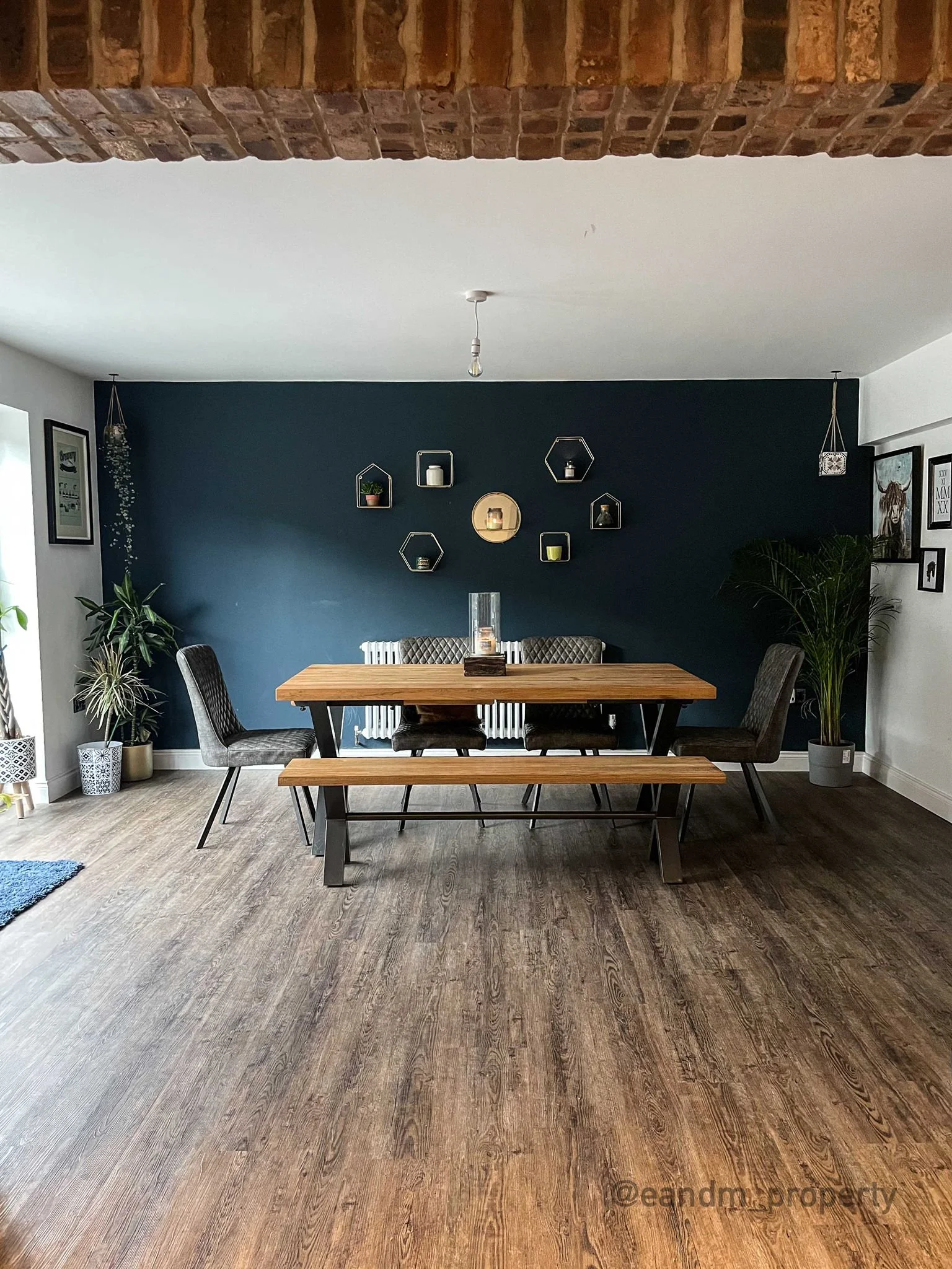 Modern dining room with wooden table, six chairs, dark accent wall with decorative shelves, plants, and framed artwork.