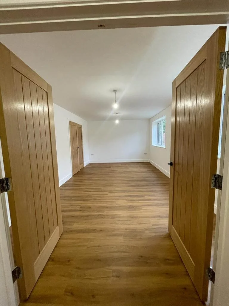 Empty room with wooden flooring, white walls, large window, and wooden double doors opening inward.