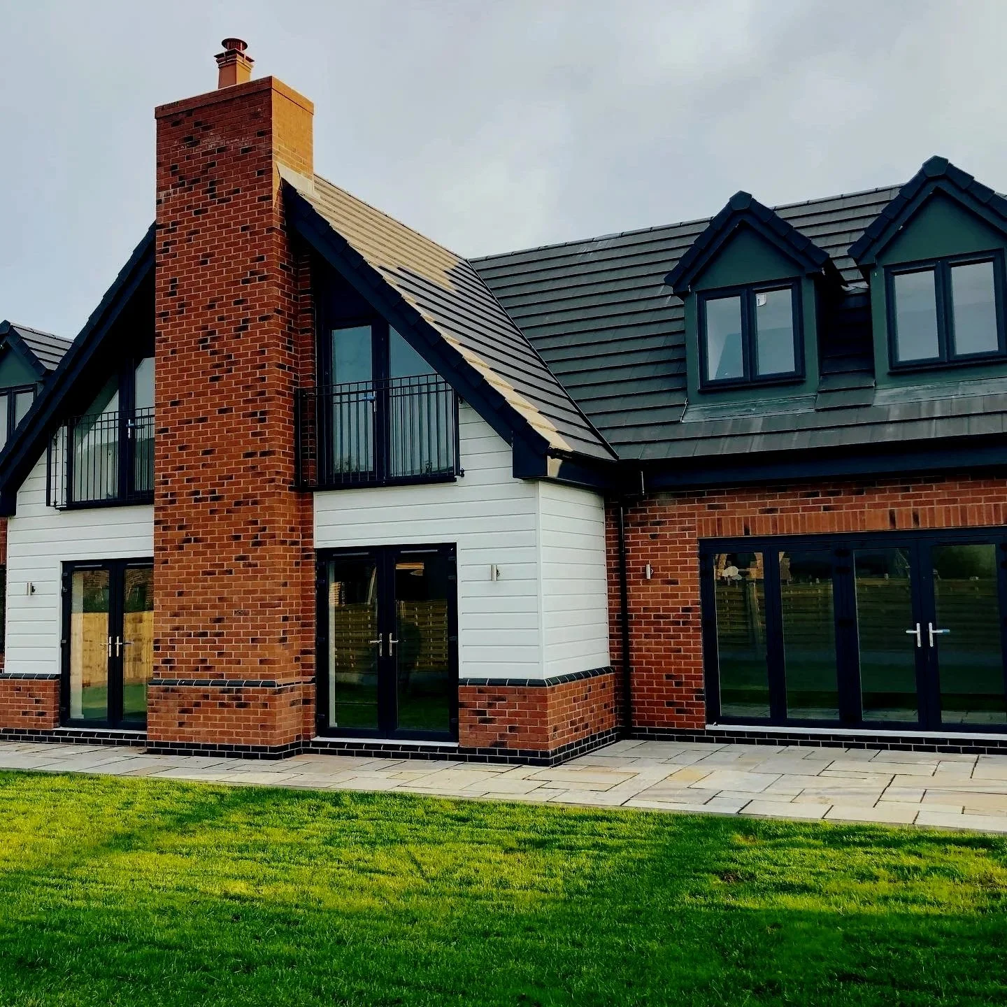Modern house with brick and white siding, large glass doors, and a patio, surrounded by a green lawn under a cloudy sky.