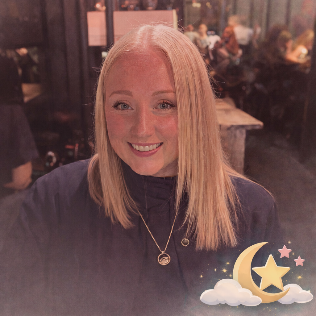 Smiling young woman with blonde hair in a dark top, sitting in a lively restaurant or bar with blurred background of other people and warm lighting, with a moon and star graphic overlay in the bottom right corner.