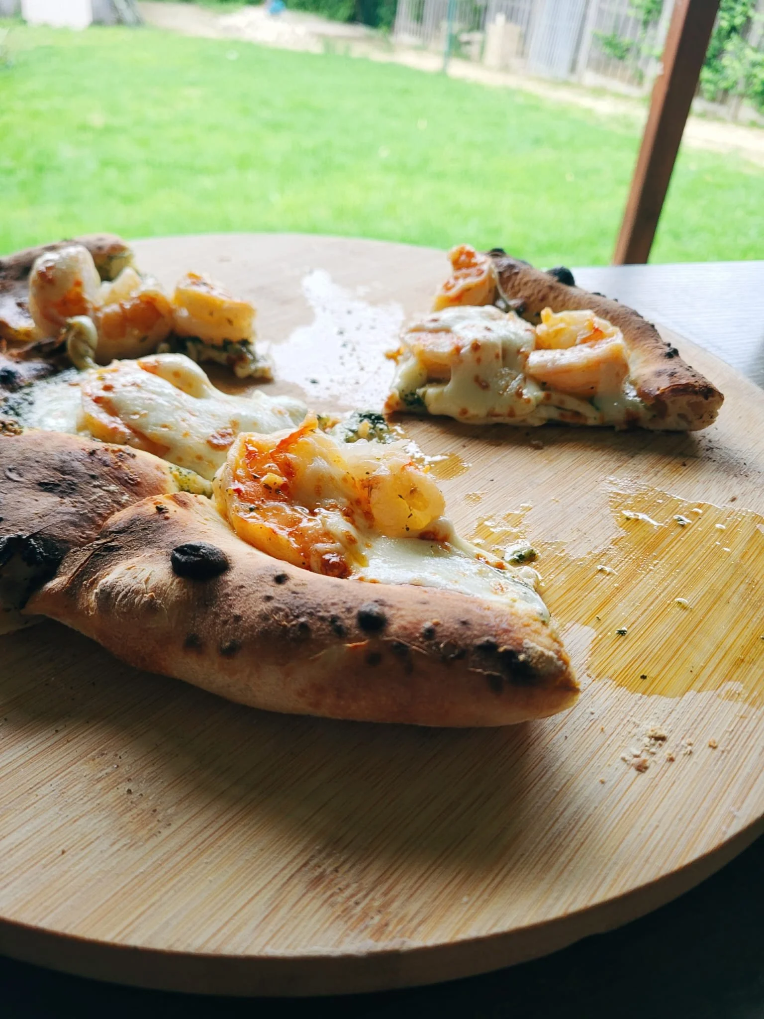 Een pizza met kaas, tomaat en kruiden, op een houten plank, met een groene tuin op de achtergrond.