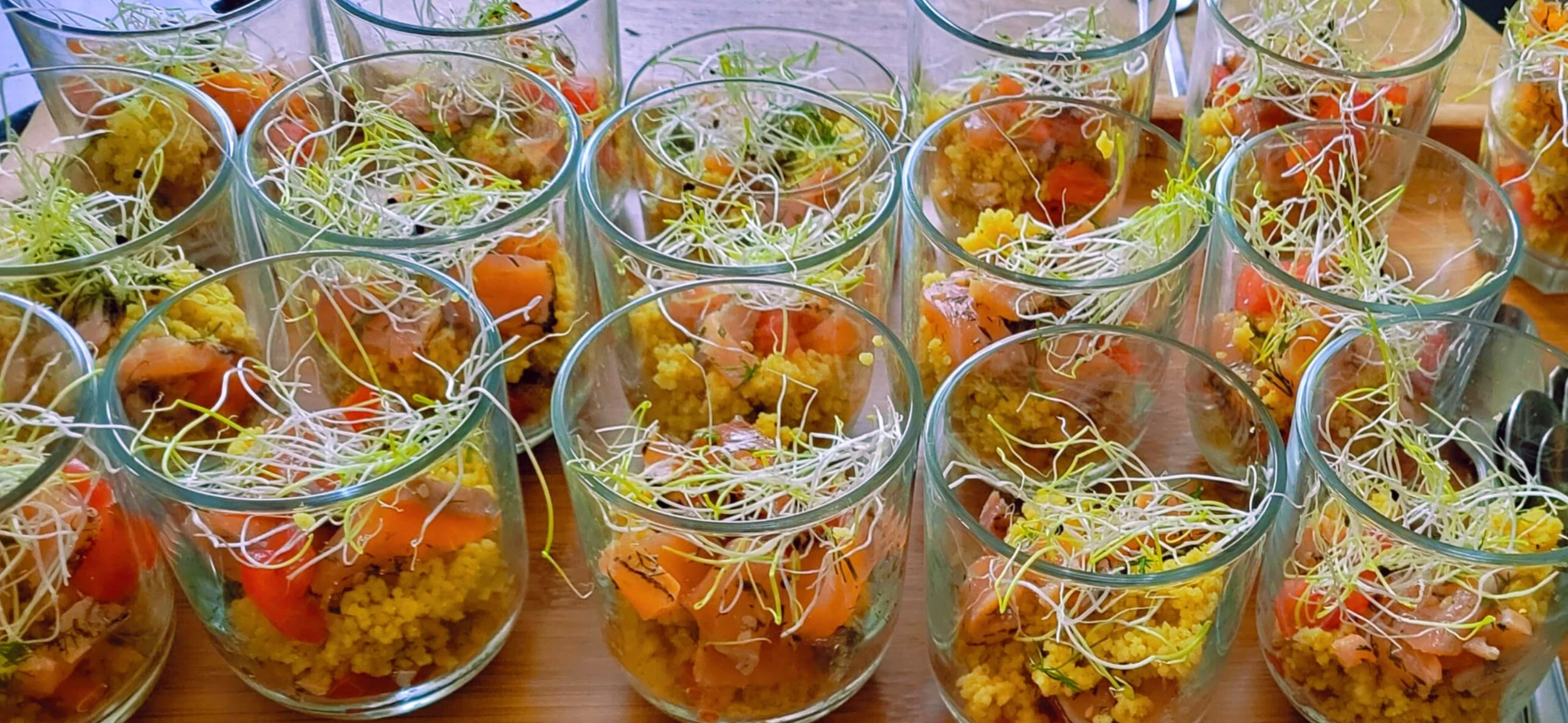 Glazen gevuld met een salade bestaande uit gekookte gele couscous, plakjes gerookte zalm en alfalfa kiemen, op een houten tafel.