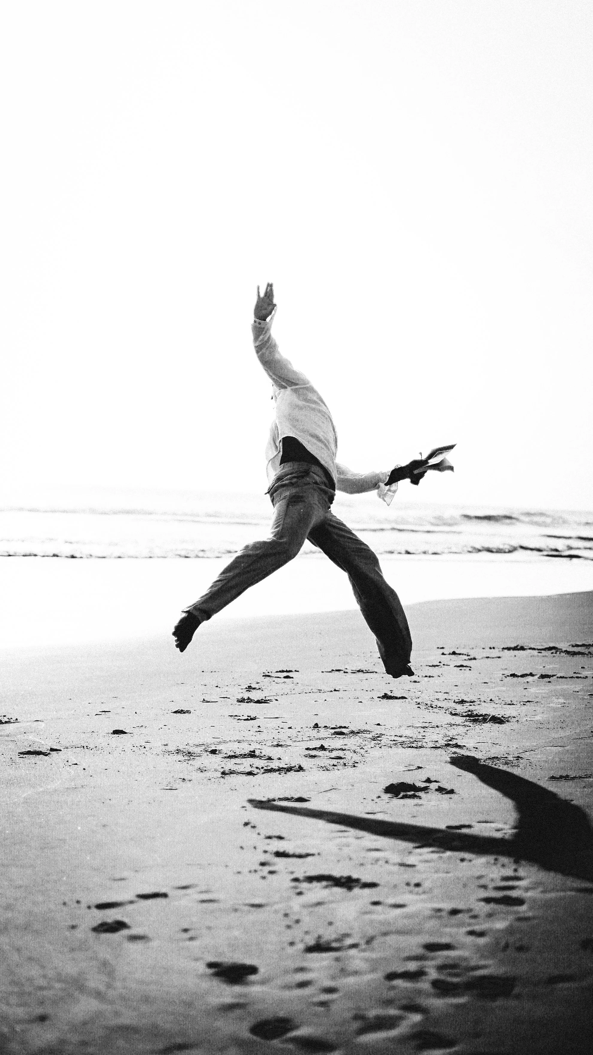 Pessoa brincando na praia, pulando com uma mochila, sombras no chão e o mar ao fundo, em preto e branco.
