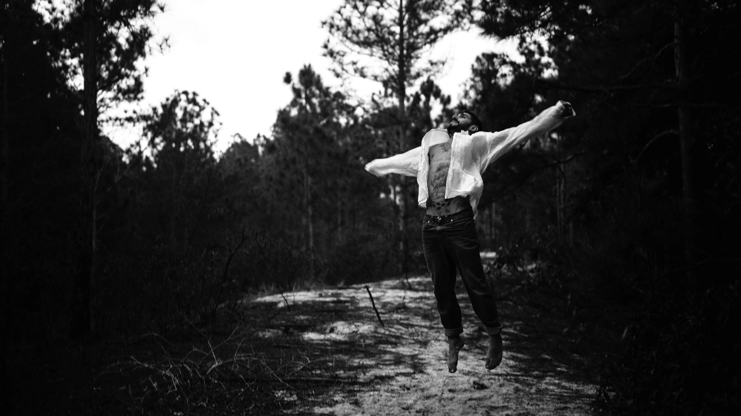 Homem sorridente pulando em uma trilha na floresta tropical, de camisa aberta e calças, ao pôr do sol ou ao amanhecer; fotografia preto e branco.