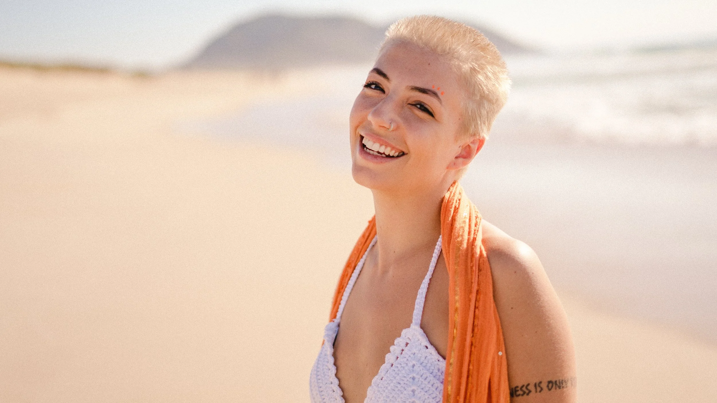 Juventude sorridente na praia com cabelo platinado, usando vestido branco de crochê e uma toalha laranja no pescoço, ao lado do mar.