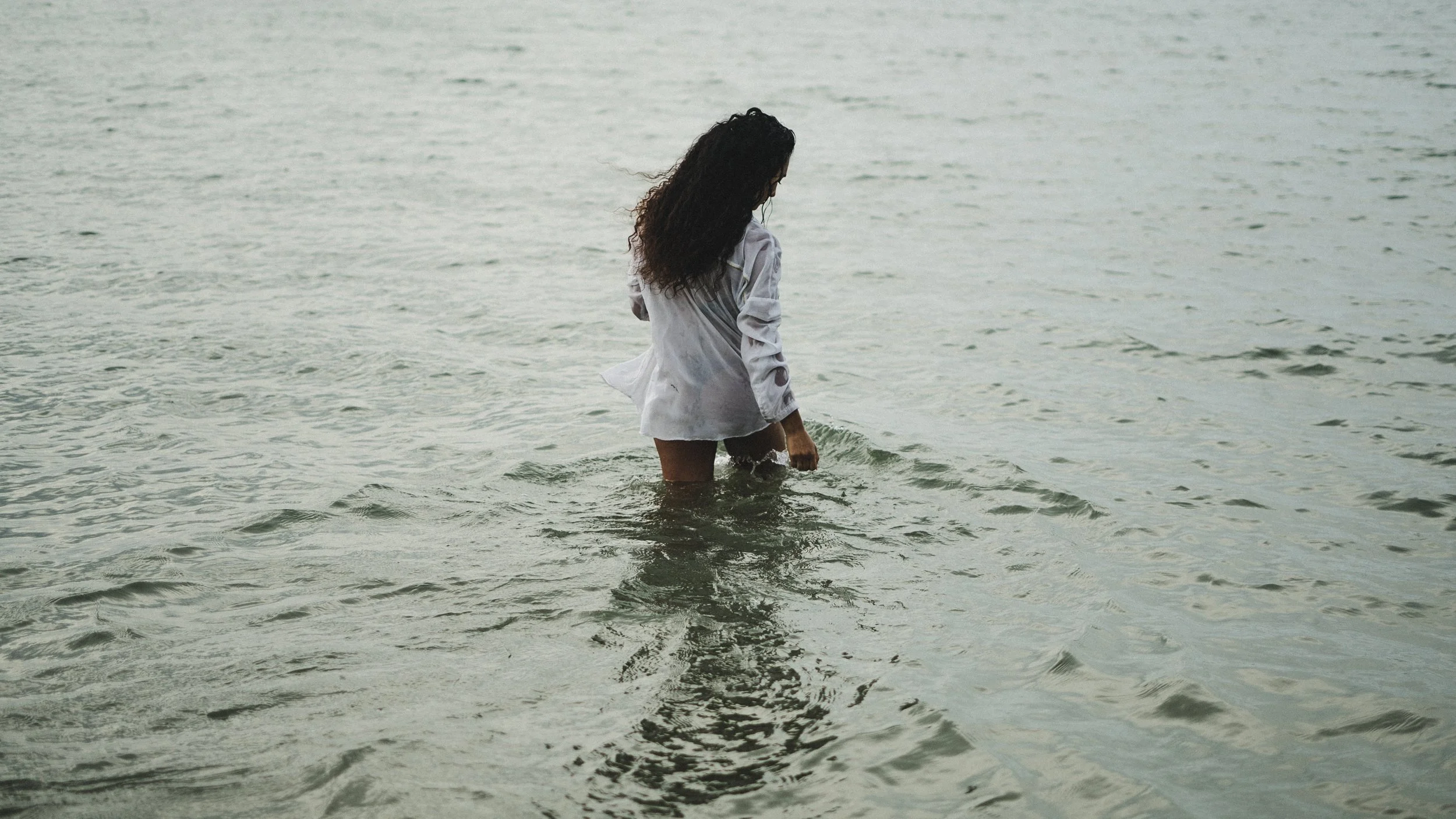 Mulher com cabelo encaracolado, vestindo camisa branca, de costas, ao pé do mar, com água até o joelho.