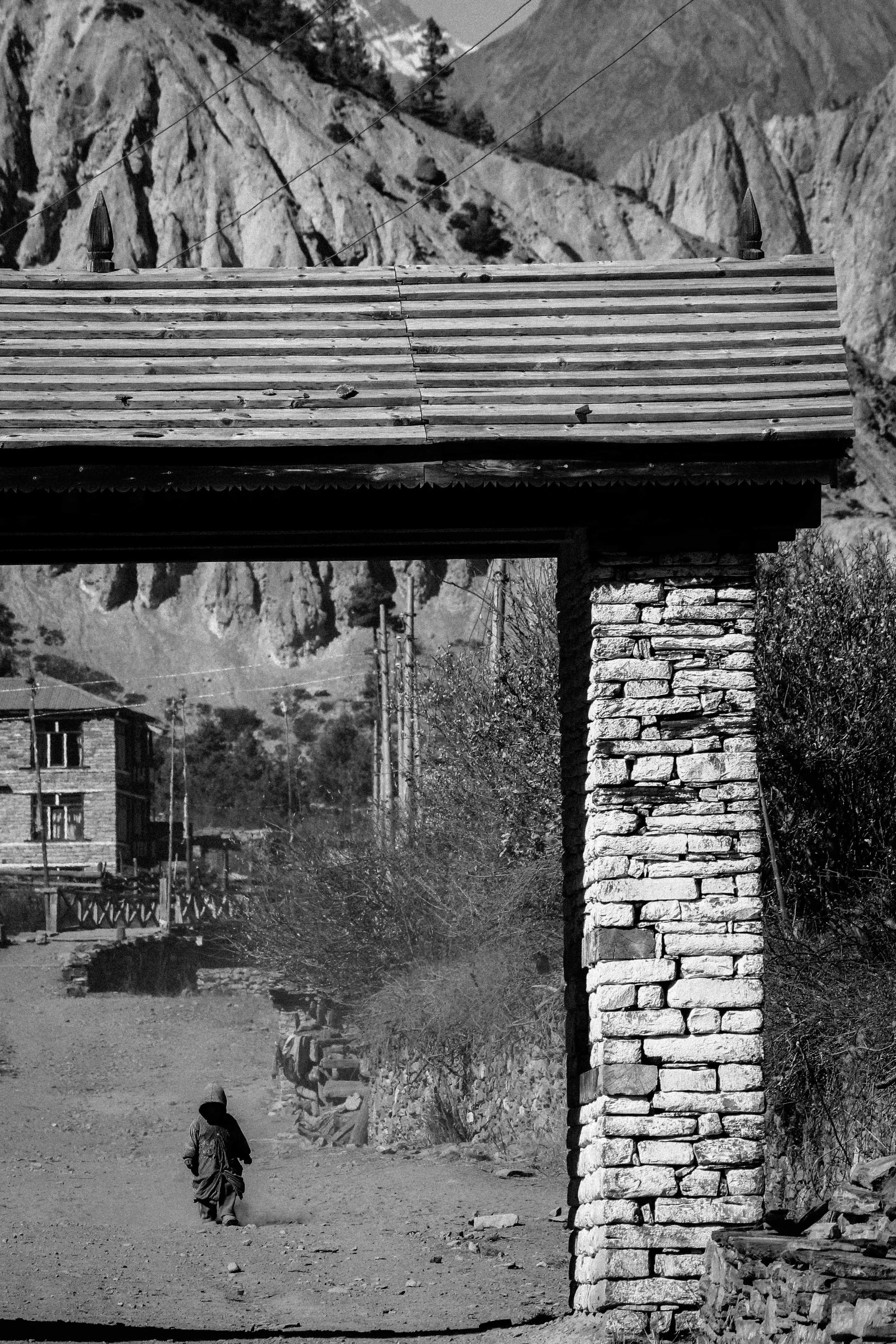 Criança caminhando por uma rua de terra em uma área rural com uma estrutura de suporte de madeira e uma coluna de pedra na frente. Montanhas ao fundo.