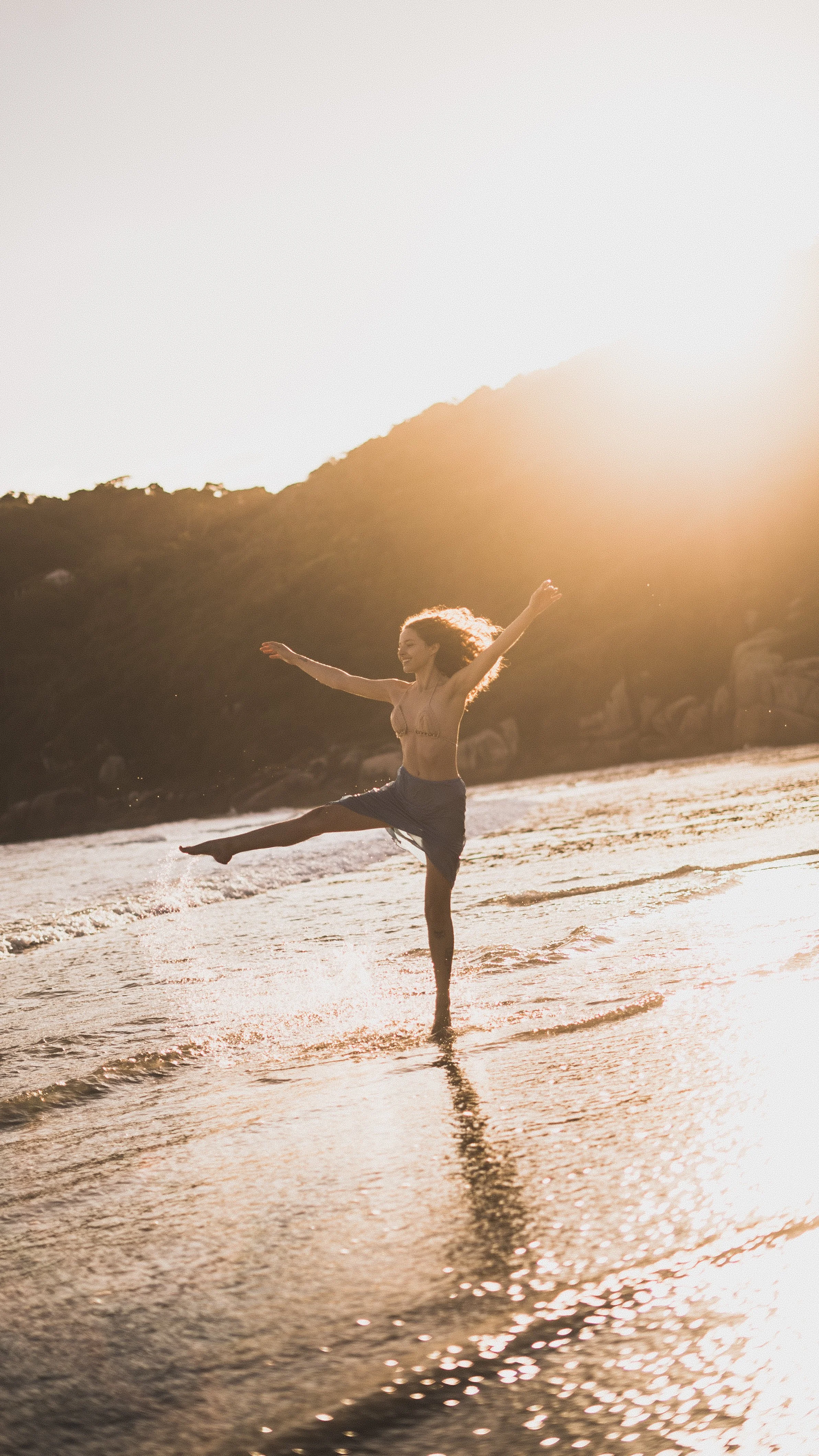 Mulher feliz dançando na praia durante o pôr-do-sol, com uma perna levantada e braços abertos.