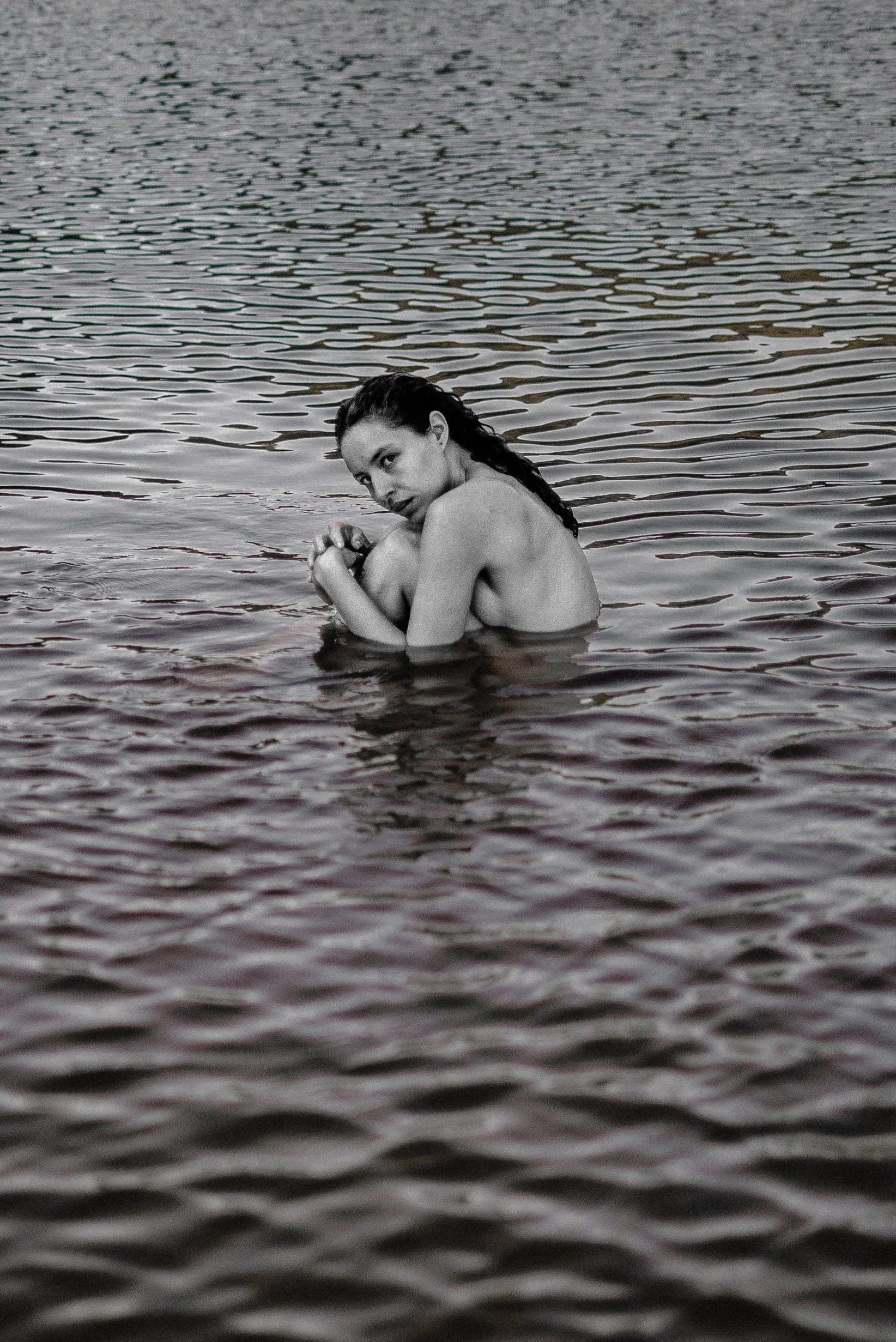 Mulher nua sentada na água de um lago, olhando para a câmera com cabelo escuro molhado e expressão pensativa.