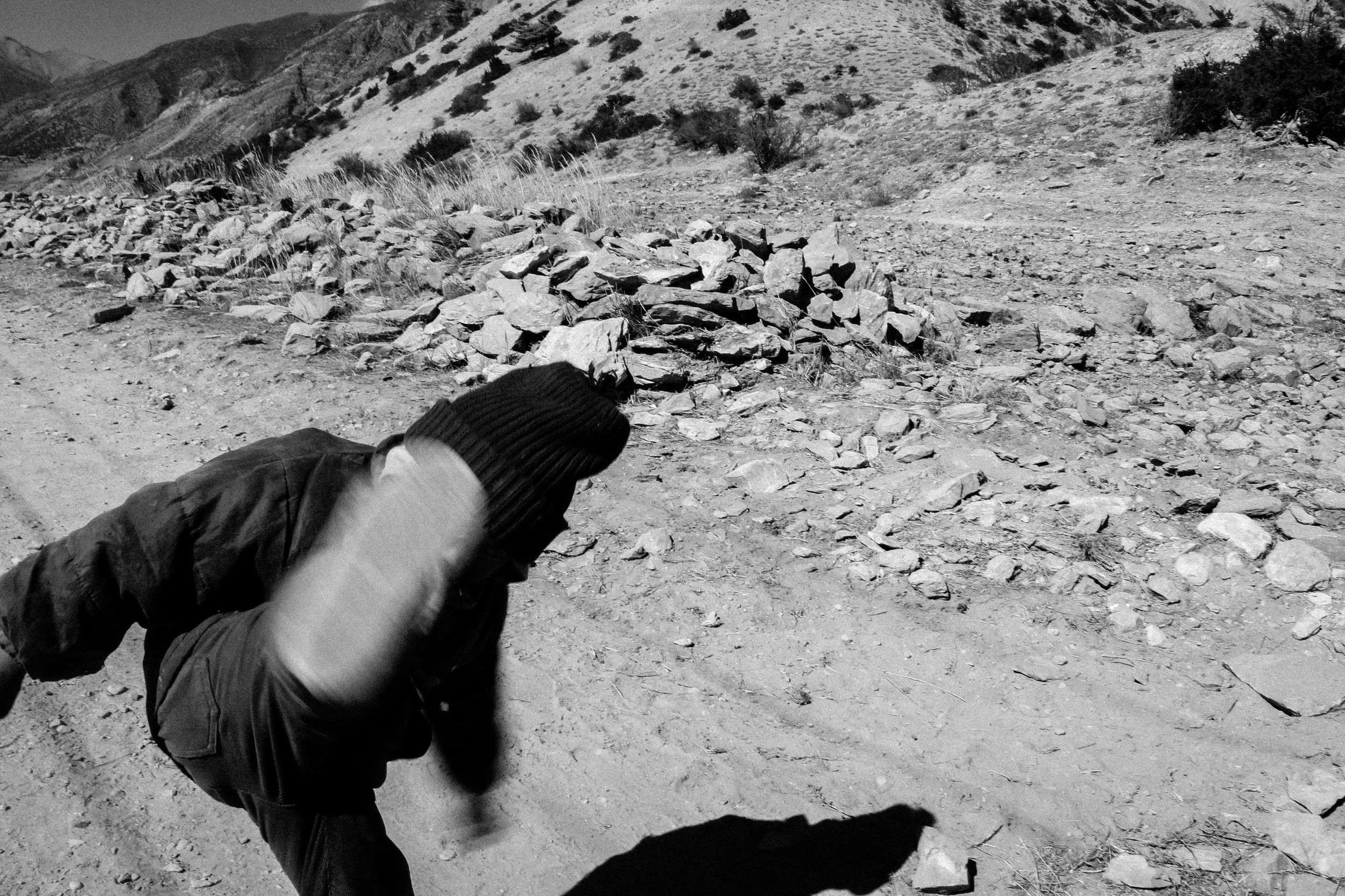 Pessoa agachada com capuz, caminhando em estrada de terra em ambiente montanhoso.