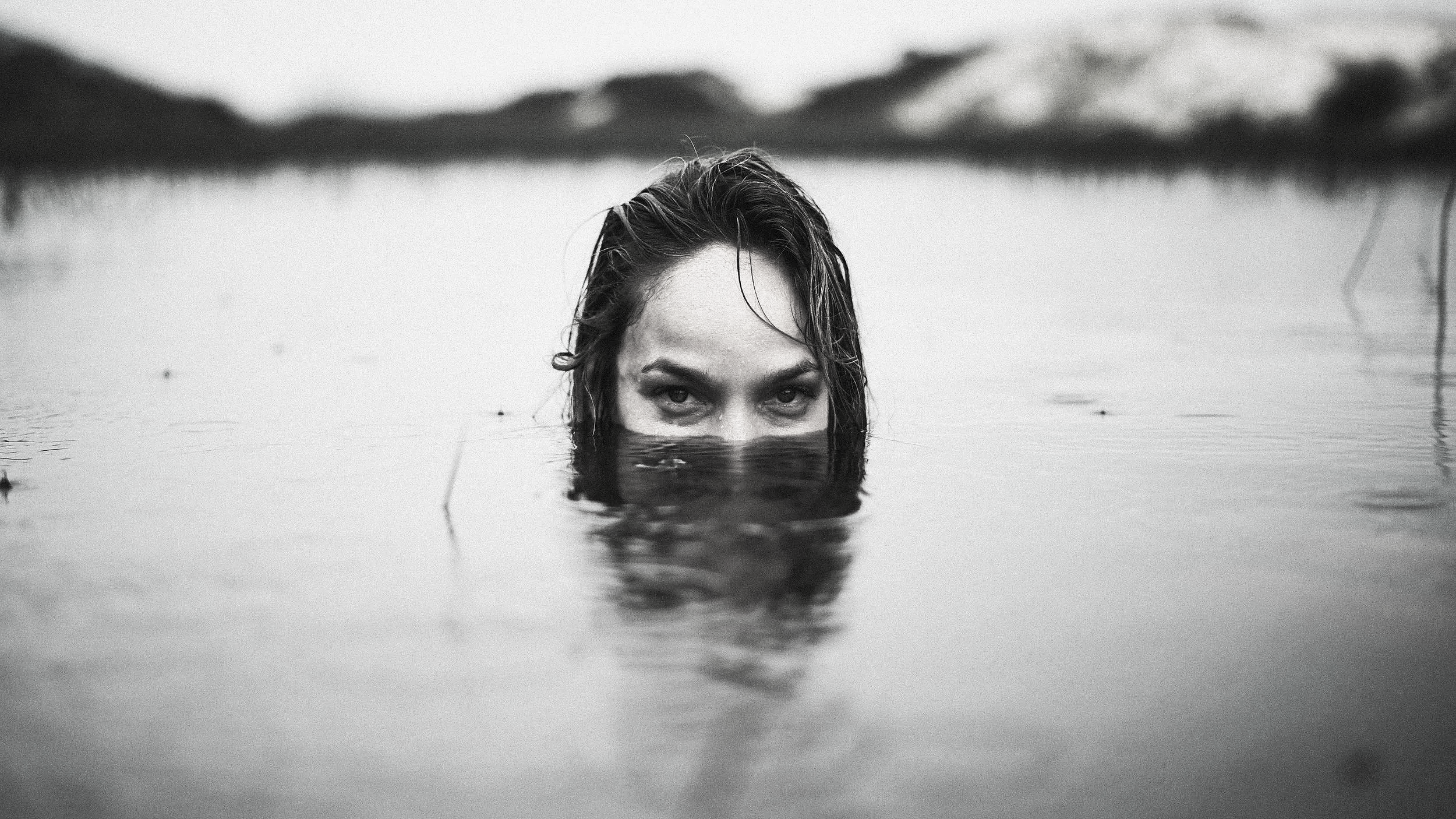 Pessoa com cabelo molhado, quase totalmente submersa na água, com apenas os olhos, sobrancelhas e parte do rosto acima da superfície da água.