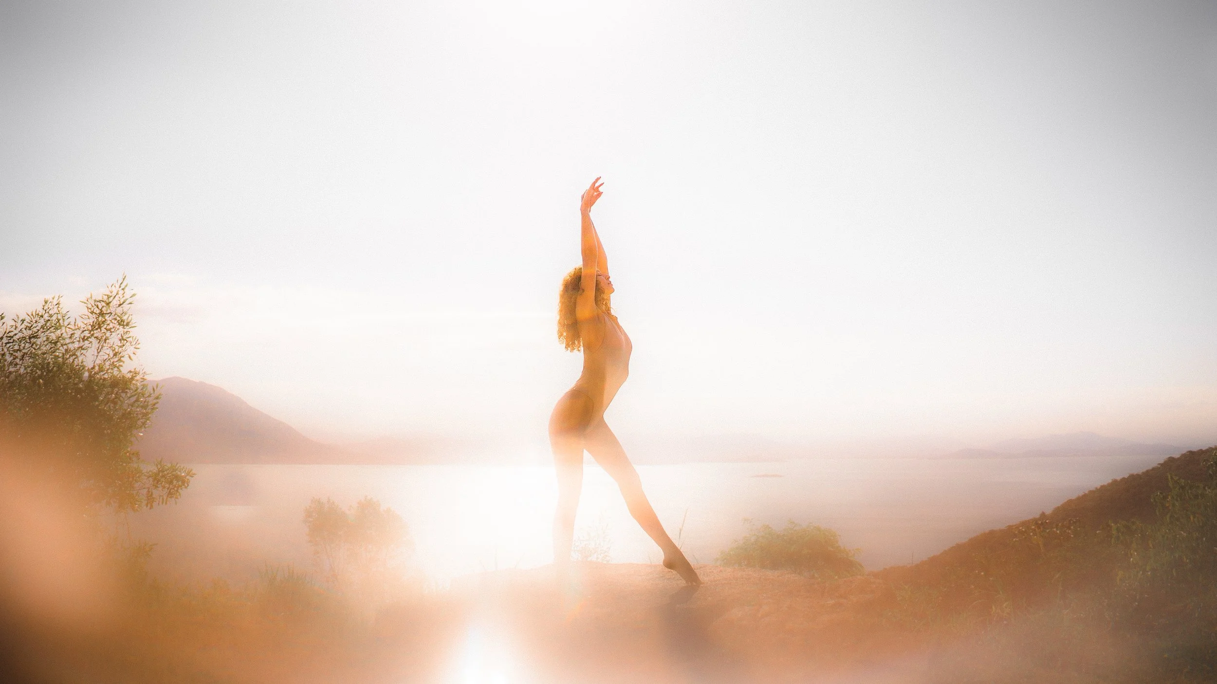 Mulher nua realizando yoga na natureza, com vista para o mar e montanhas ao fundo, durante o pôr do sol.