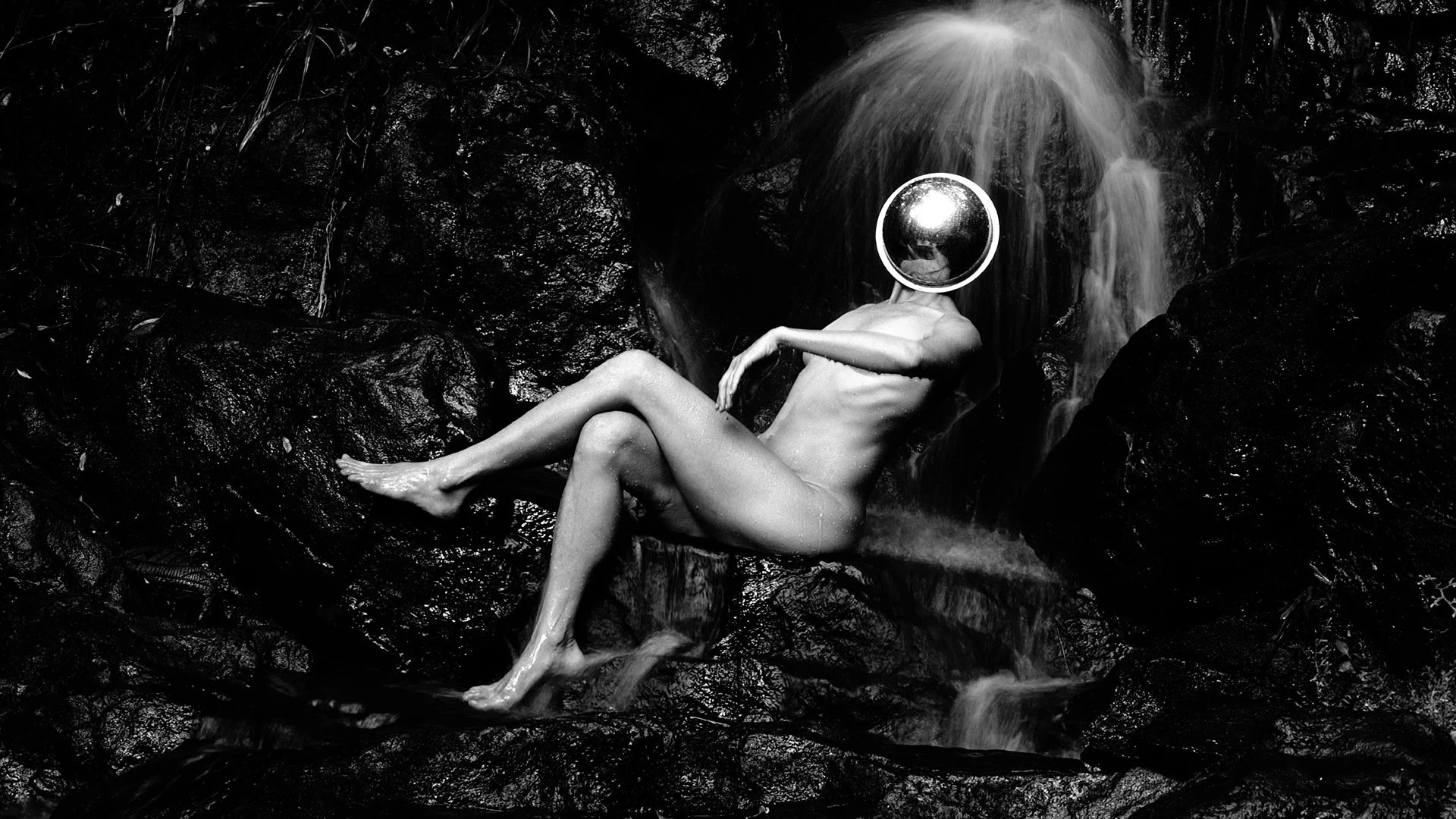 Figura humana nua com cabeça de espelho, sentada em rochas ao lado de uma cachoeira, em ambiente de natureza, em uma imagem em preto e branco.