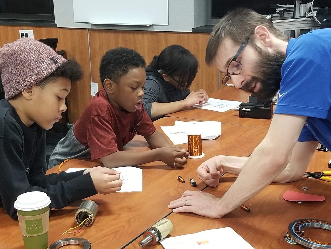 A group of three children and a man with glasses working on a science project at a table with various electronic components and papers.