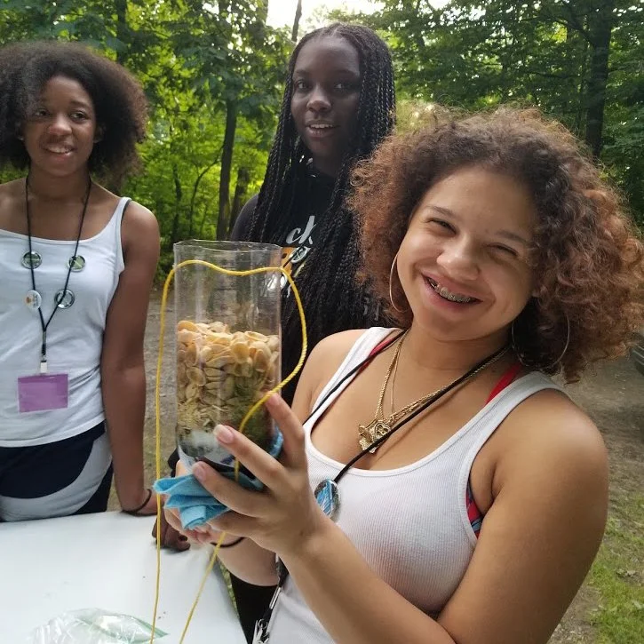 Three women outdoors in a wooded area, one smiling woman holding a cylindrical container filled with pasta and a yellow string attached, other two women standing beside her.