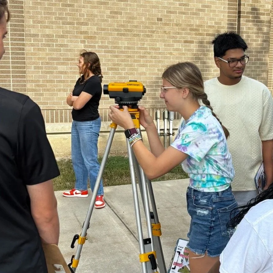A young woman using a total station surveying instrument outdoors, surrounded by a few people, with a brick building in the background.